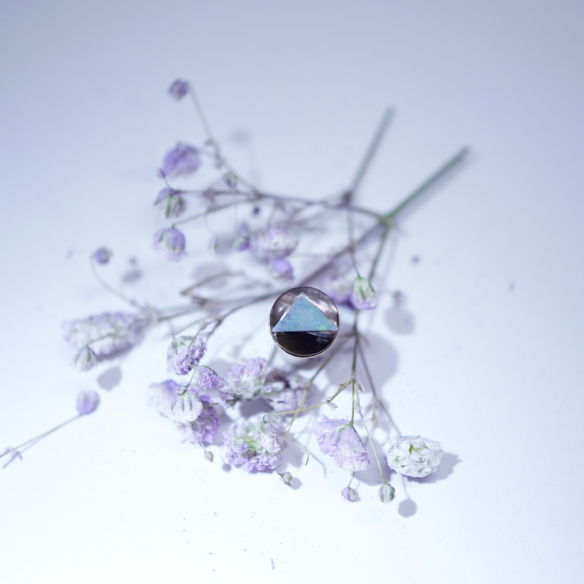 A single round stud earring with a faceted iridescent blue-green gemstone in a dark bezel setting, artistically placed among dried lavender and white baby's breath flowers on a light, cool-toned background.