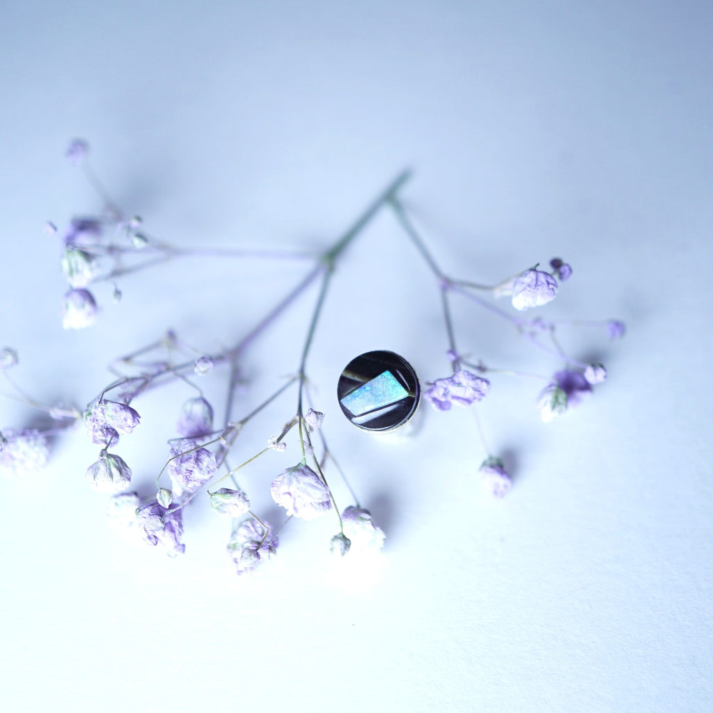 A silver ring with a rectangular iridescent blue-green stone, styled on delicate dried lavender sprigs against a pale blue background.