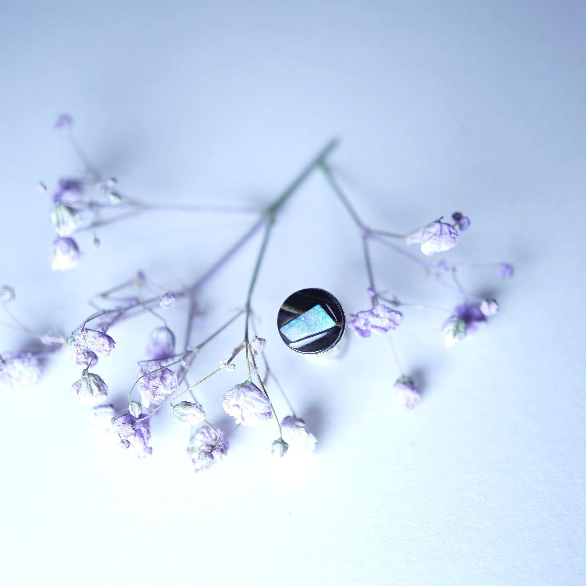 A silver ring with a rectangular iridescent blue-green stone, styled on delicate dried lavender sprigs against a pale blue background.