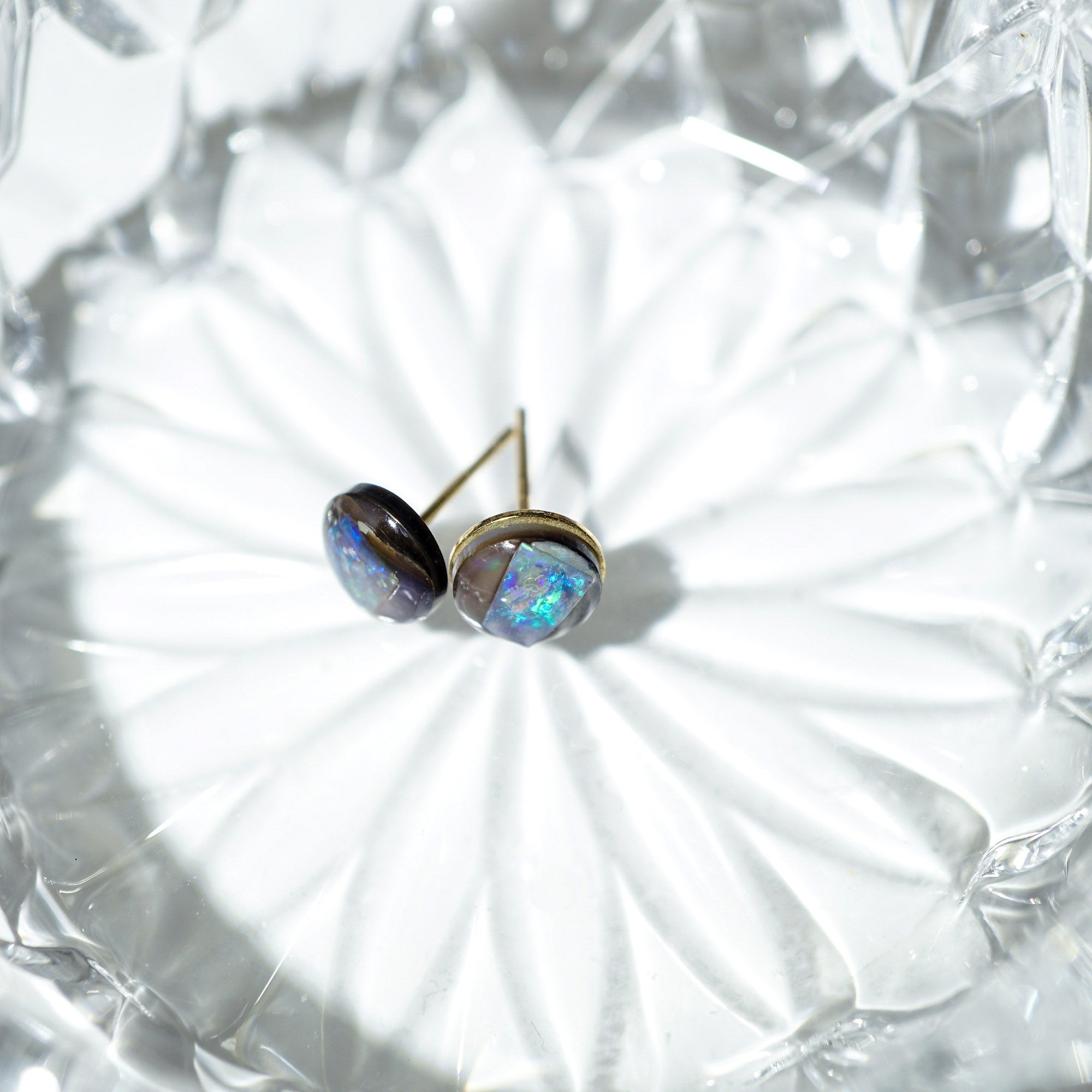 A pair of oval stud earrings with iridescent opal-like stones in gold-toned settings, resting on a white textured glass dish.