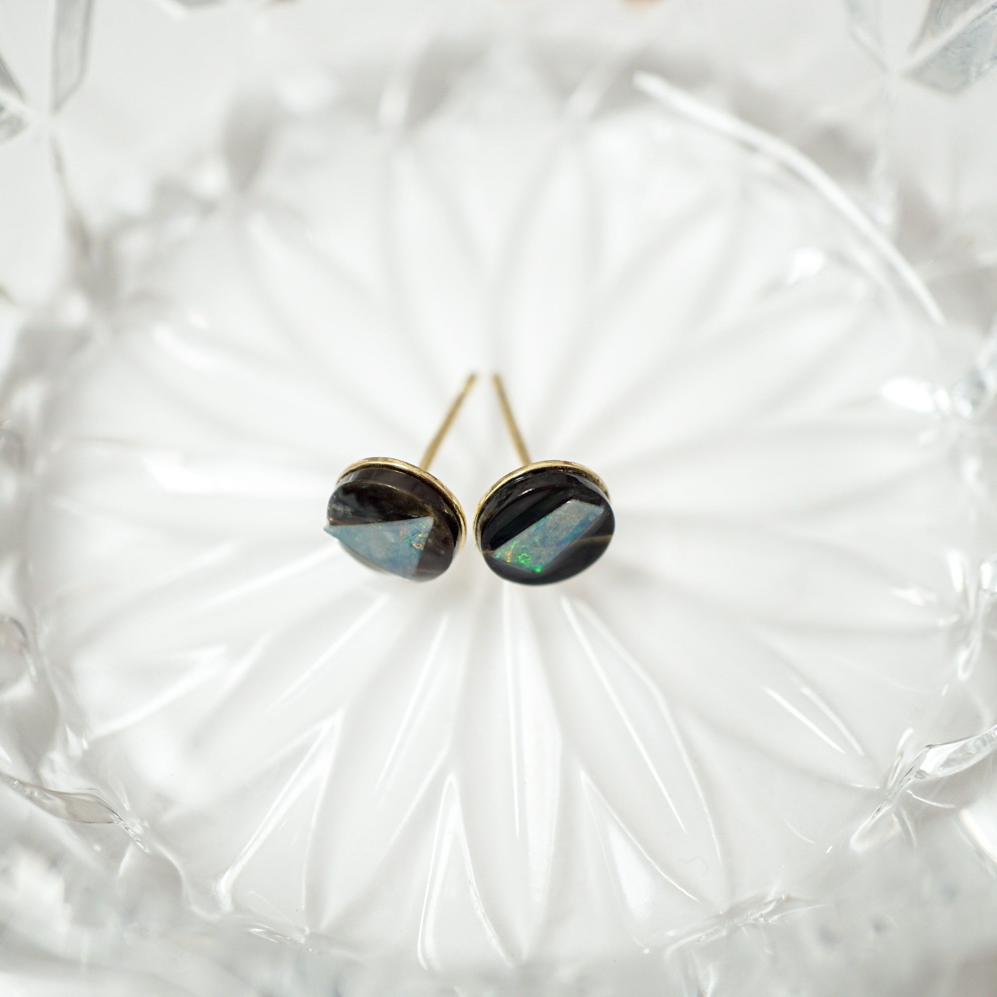 A pair of opal stud earrings with dark iridescent opal-like centers and gold-toned settings, resting on a clear faceted glass dish.
