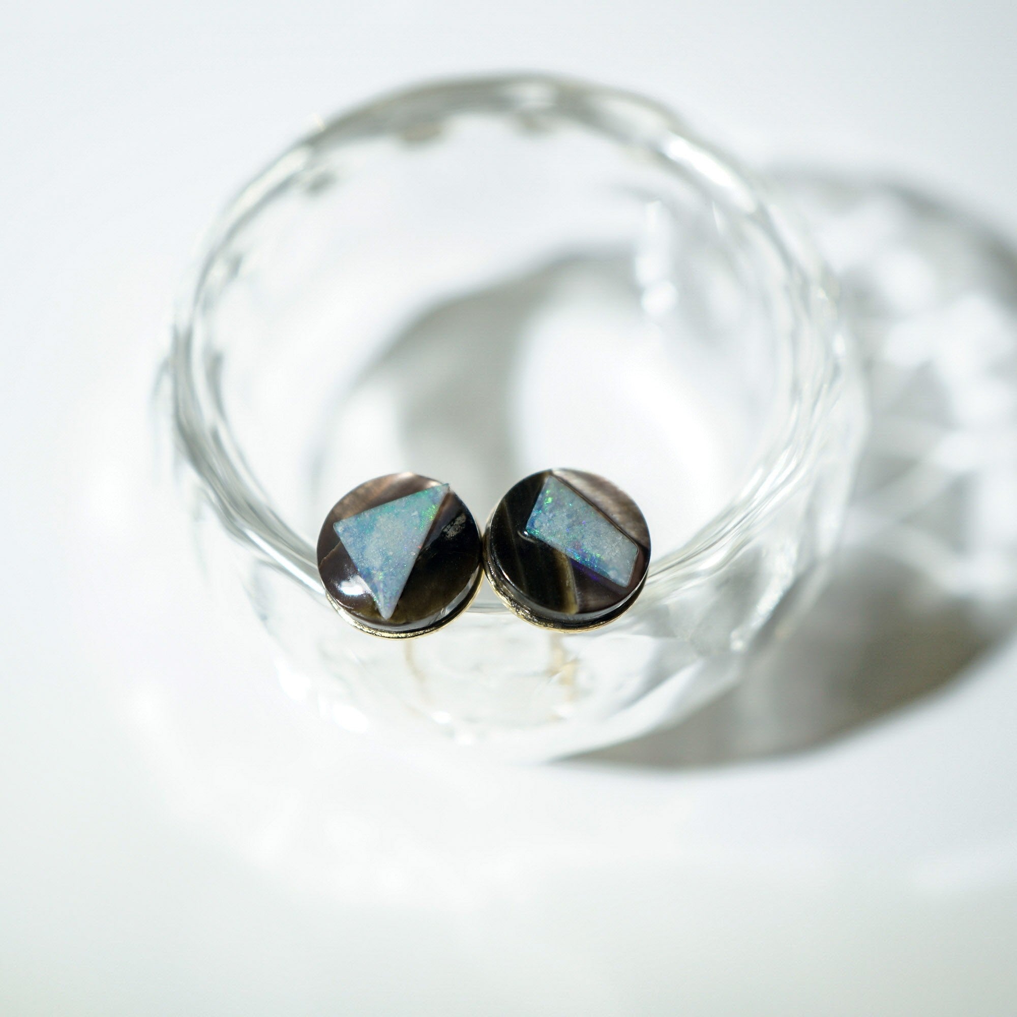 A pair of round stud opal earrings with faceted iridescent blue and black triangular stones in silver bezels, resting inside a clear glass dish on a white surface.