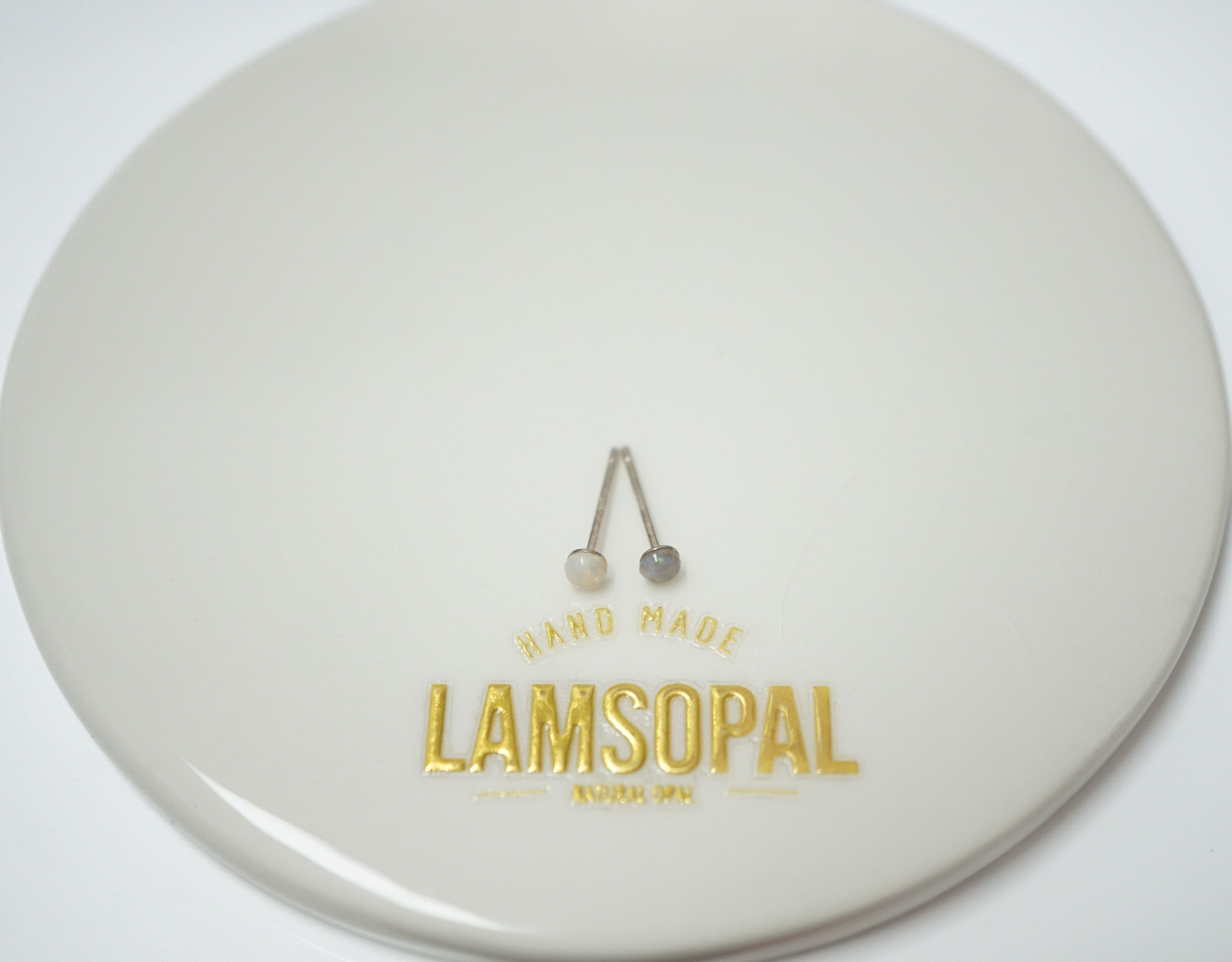 A pair of silver-colored barbell stud earrings displayed on a glossy white circular plate with gold foil branding that reads 'HAND MADE LAMSOPAL'.