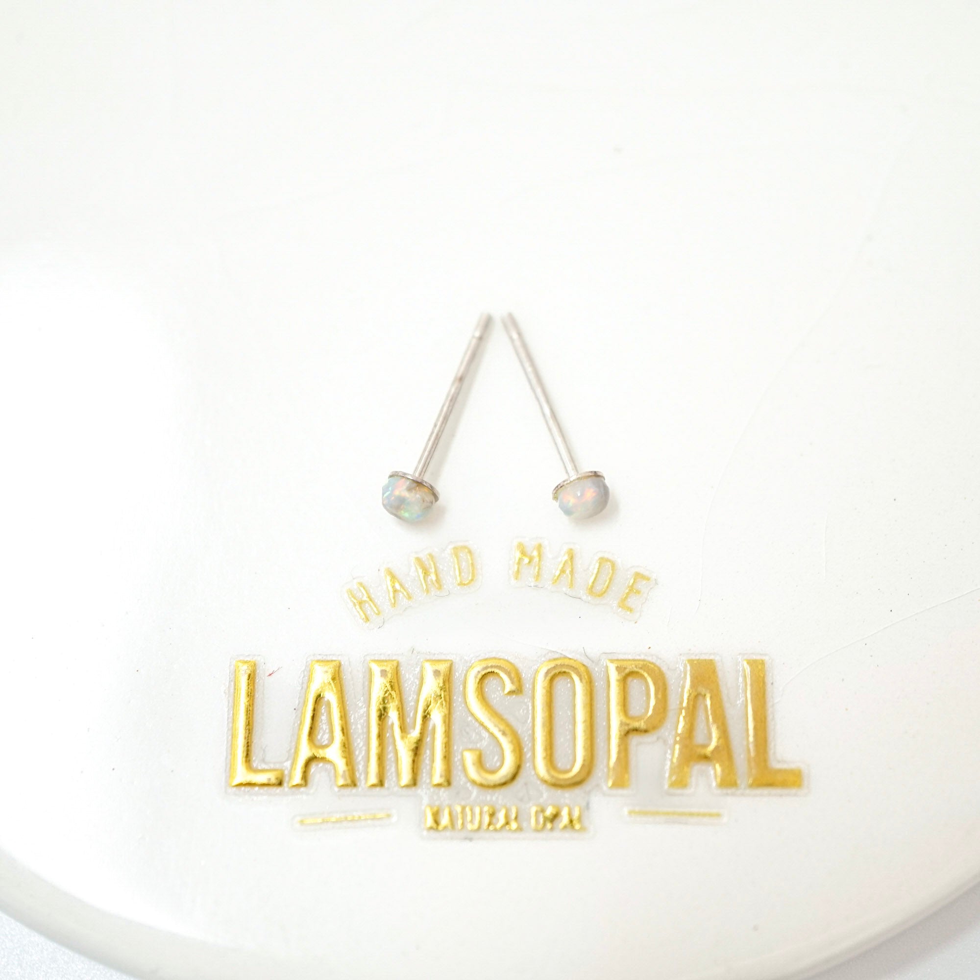 A pair of silver-toned stud earrings with round, iridescent opal-like beads, displayed above a gold foil logo that reads 'HAND MADE LAMSOPAL NATURAL DEG'.