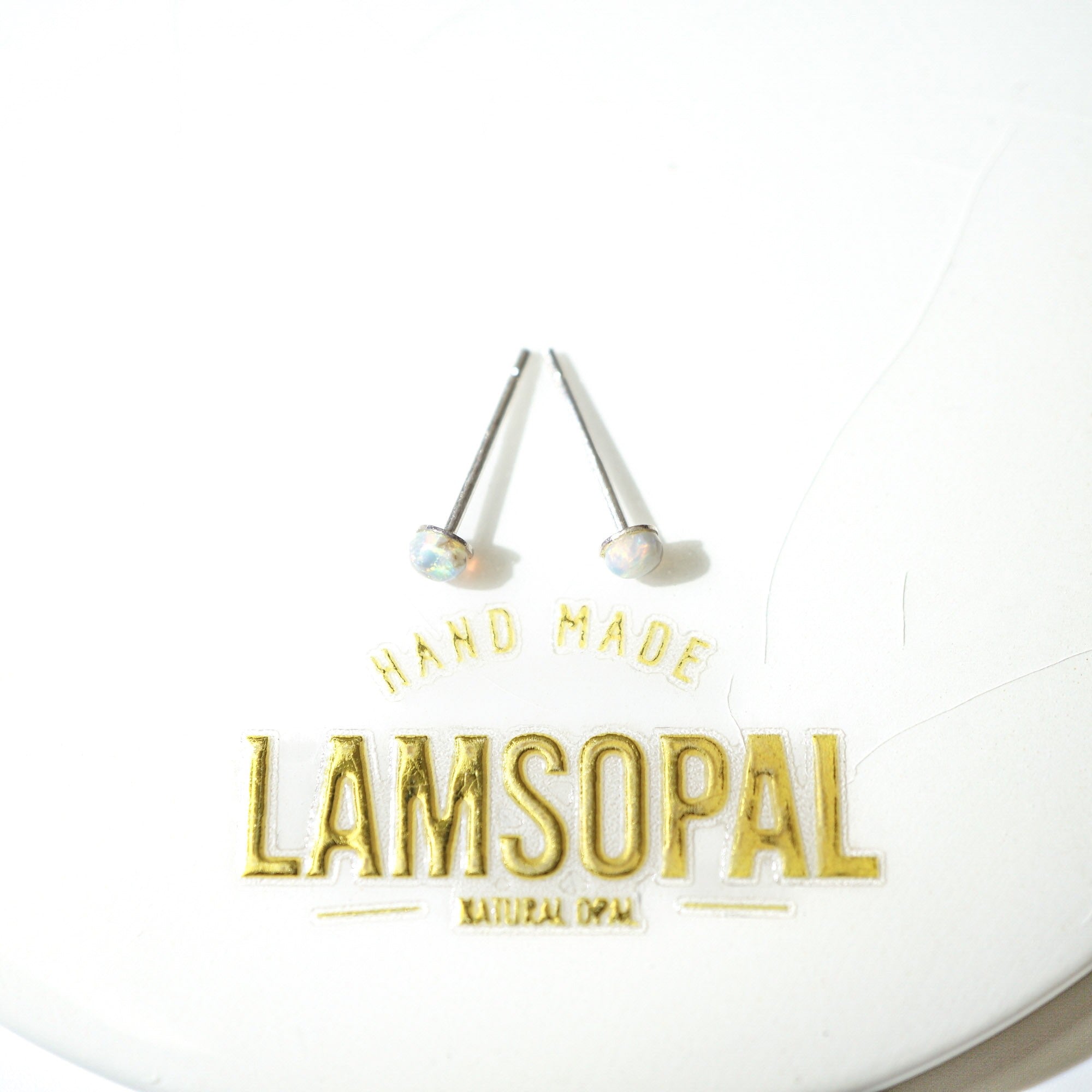 A pair of Lamsopal natural opal stud earrings with silver posts and round iridescent opal gemstones, displayed on a white circular backing card with gold foil branding.