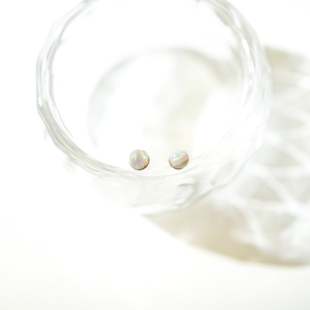 Two small iridescent oval stud earrings resting inside a clear glass dish on a bright white background.