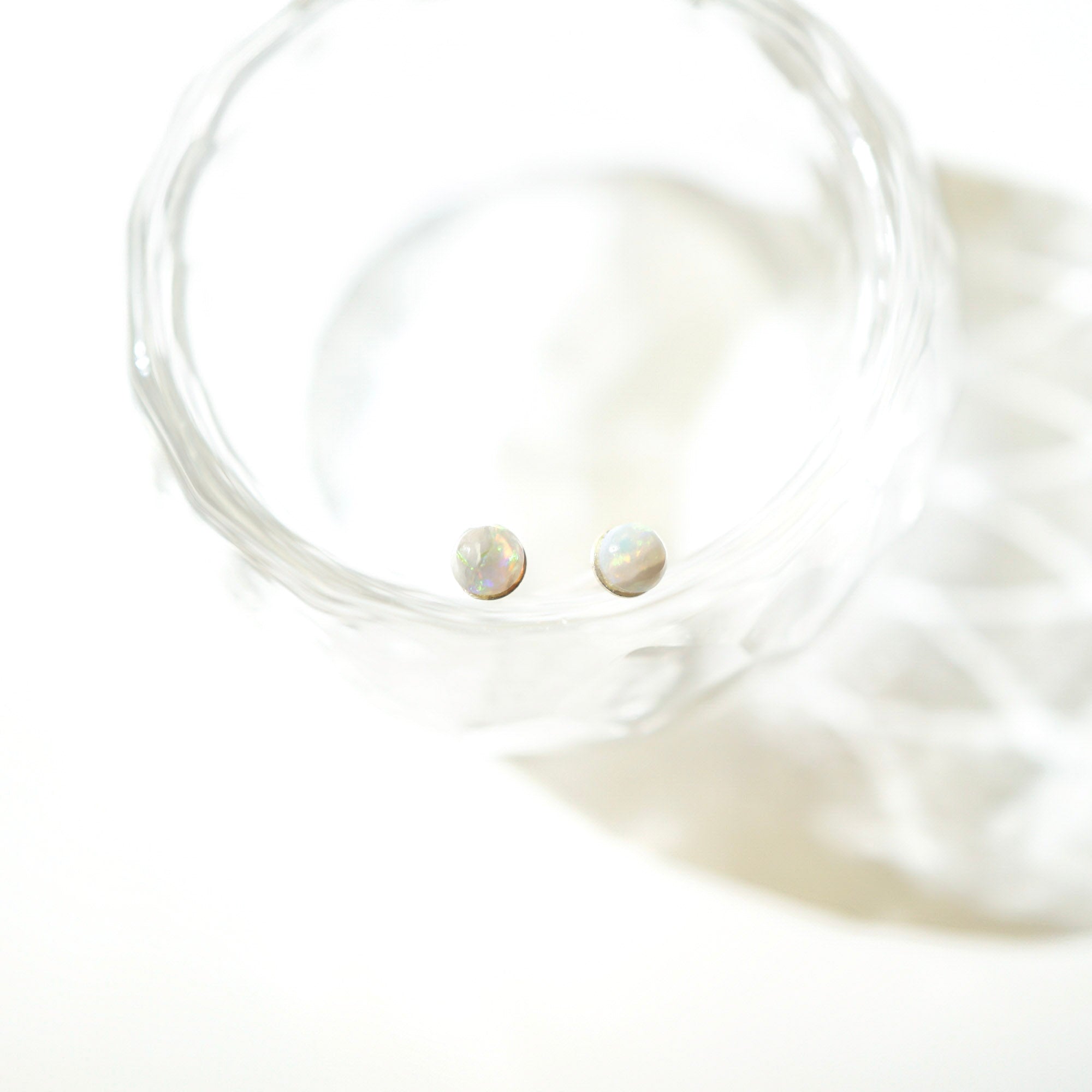 Two small iridescent oval stud earrings resting inside a clear glass dish on a bright white background.