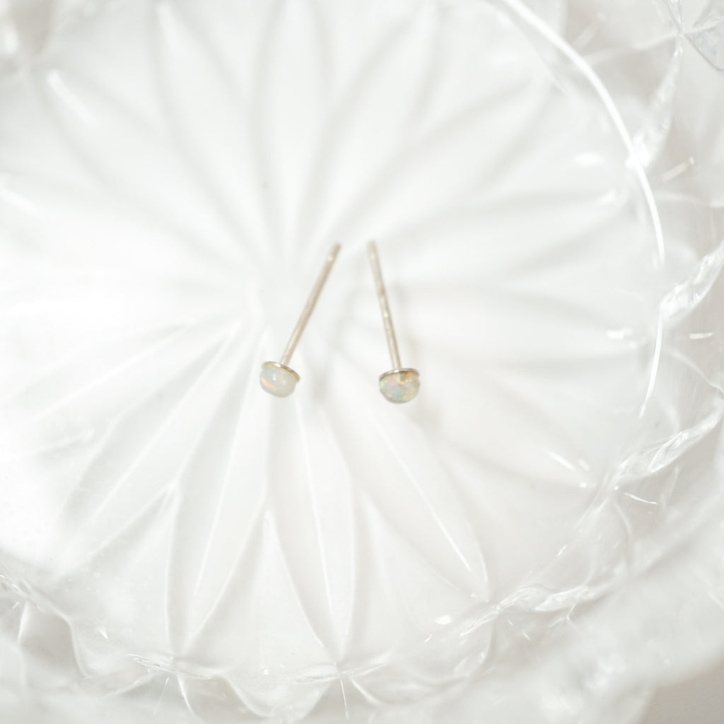 A pair of delicate gold stud earrings with small, oval-shaped iridescent opal gemstones, resting on a textured white glass plate.