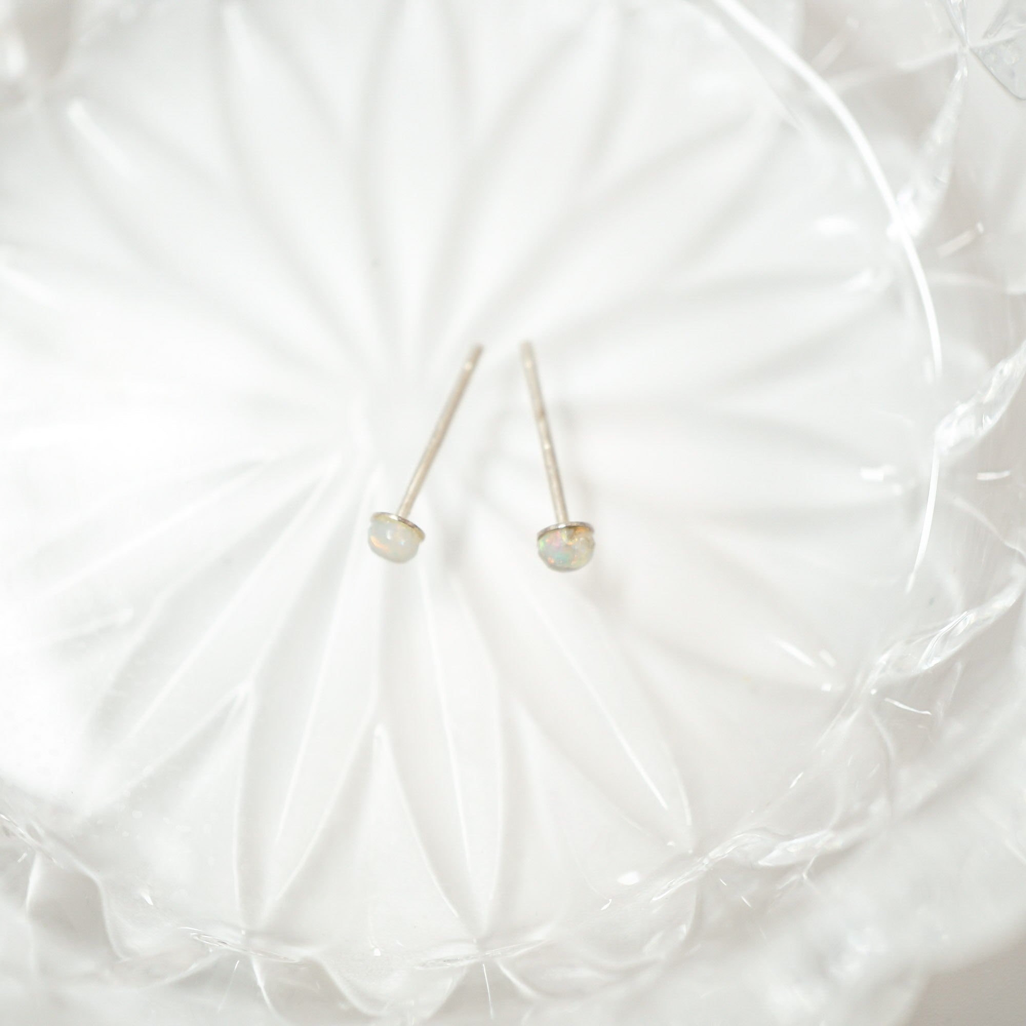 A pair of delicate gold stud earrings with small, oval-shaped iridescent opal gemstones, resting on a textured white glass plate.