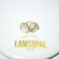 A pair of gold stud earrings with round iridescent cabochon gemstones, displayed on a white surface above a gold foil brand label reading 'LAMSOPAL'.
