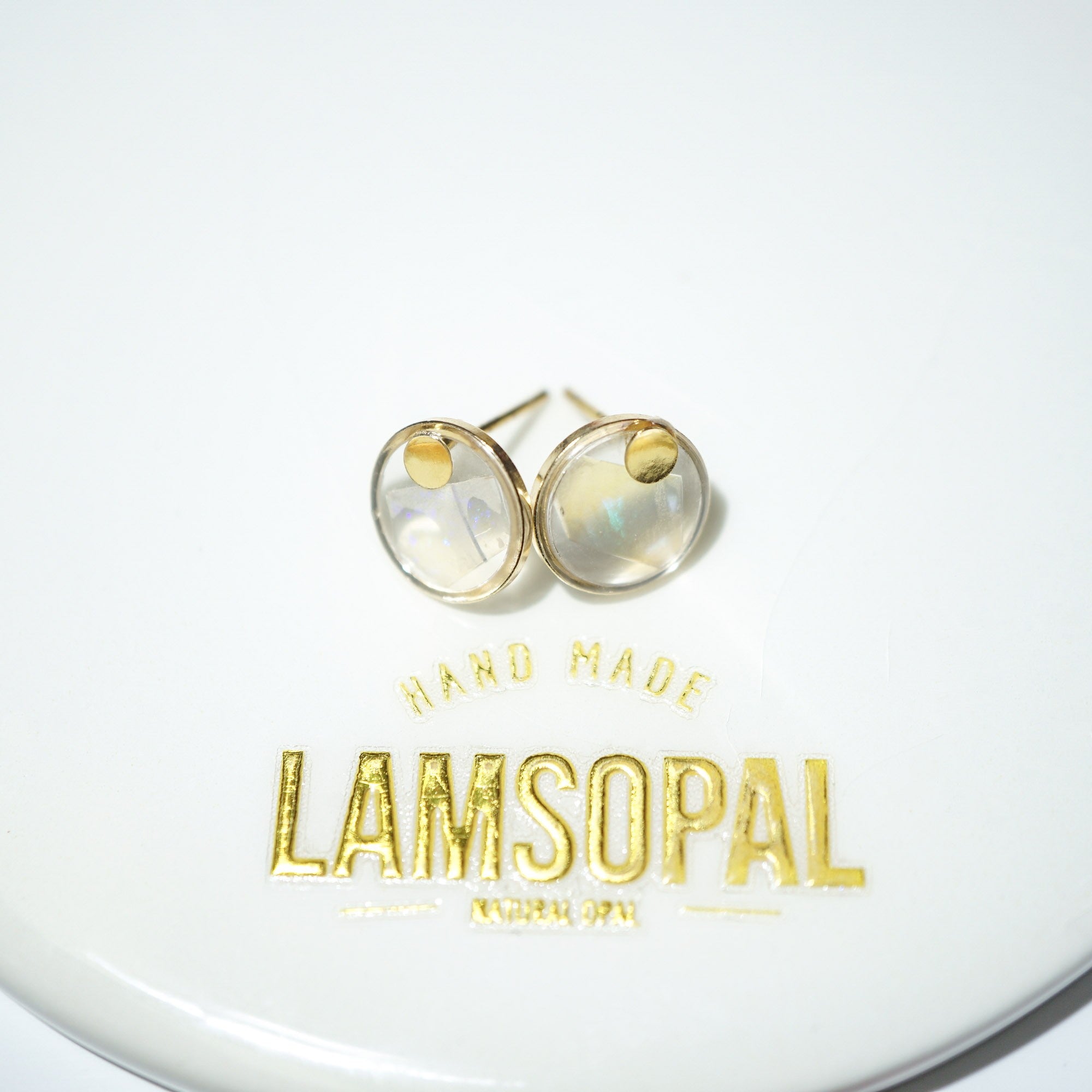 A pair of gold stud earrings with round iridescent cabochon gemstones, displayed on a white surface above a gold foil brand label reading 'LAMSOPAL'.