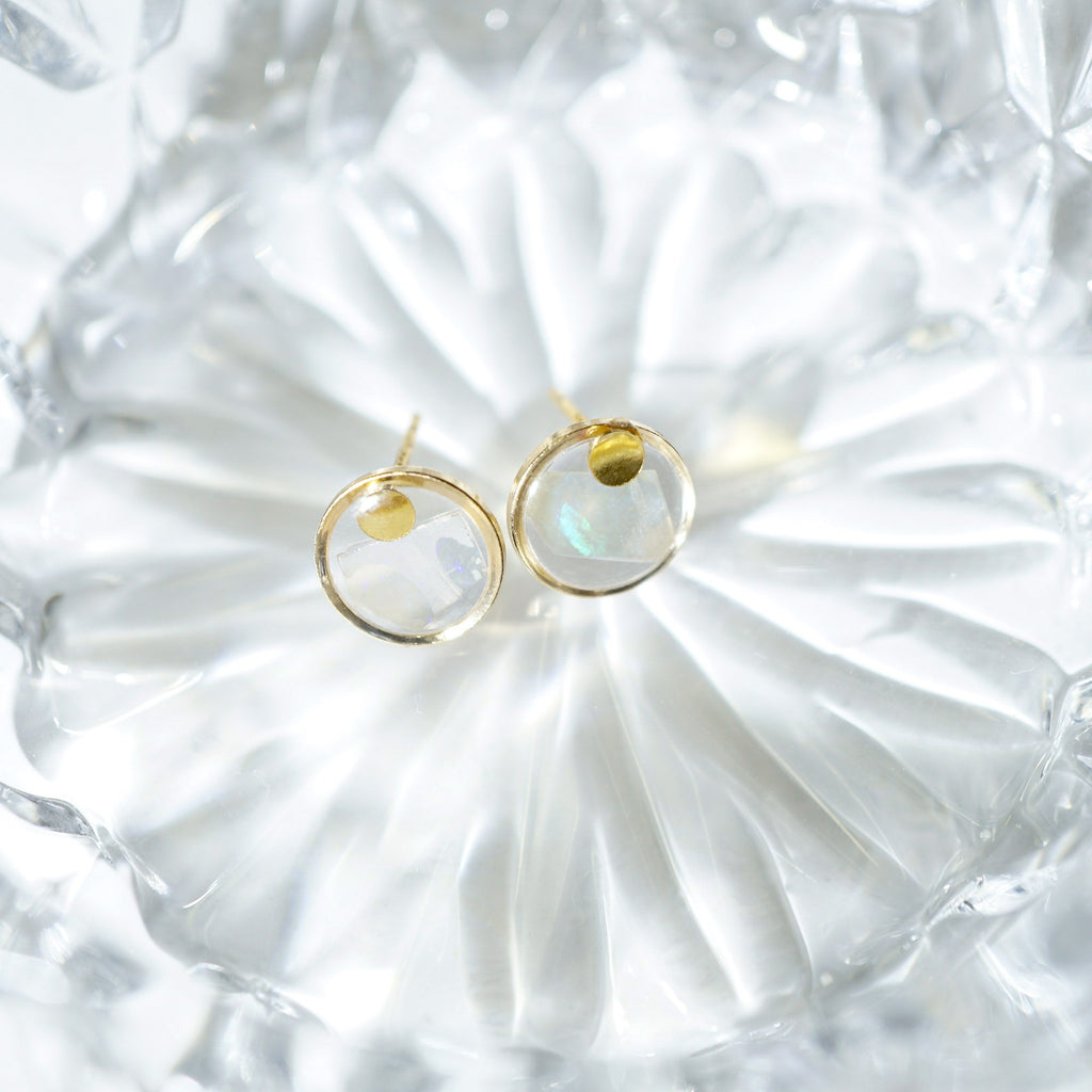 A pair of gold hoop earrings with iridescent white crescent moon charms, displayed on a textured clear glass dish.