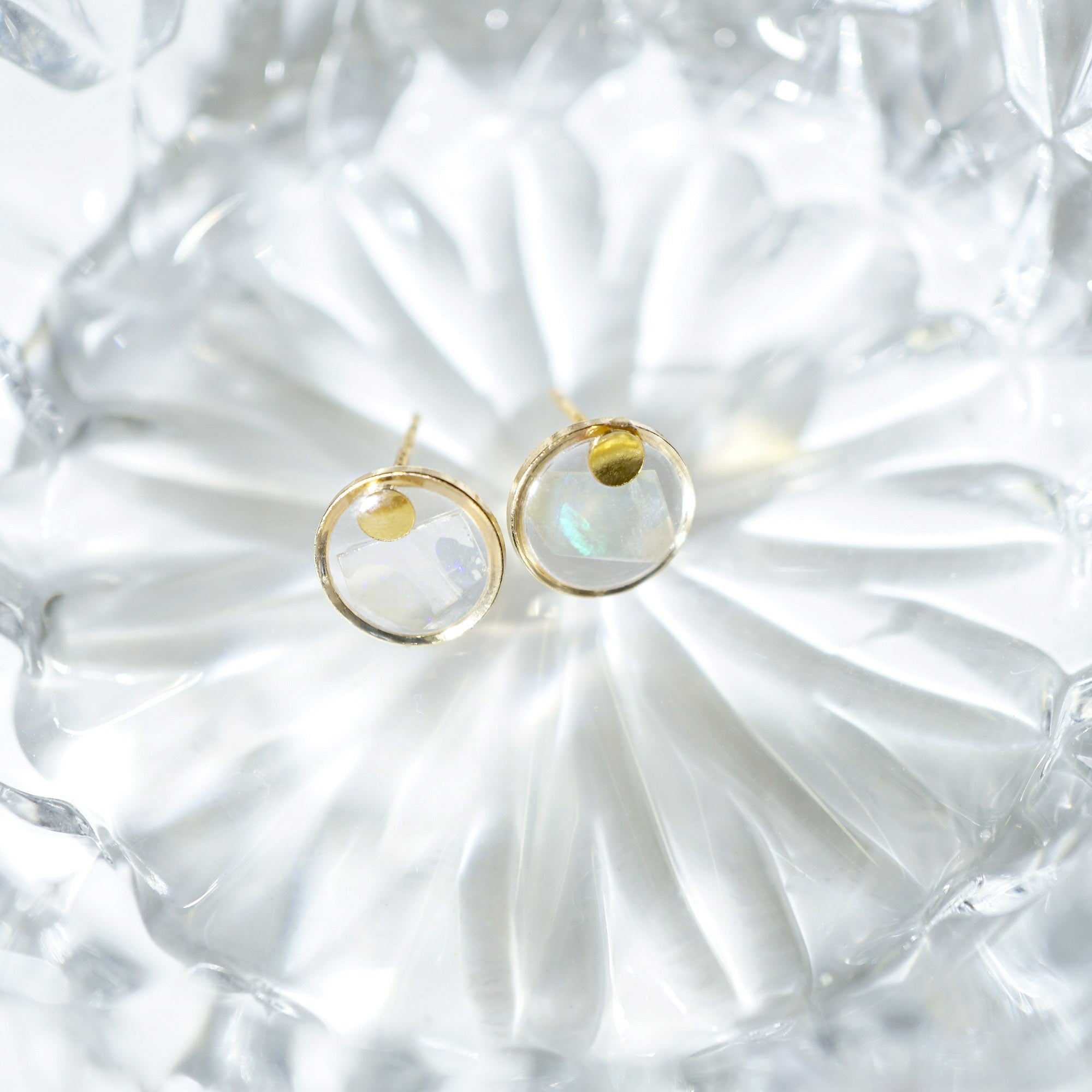 A pair of gold hoop earrings with iridescent white crescent moon charms, displayed on a textured clear glass dish.