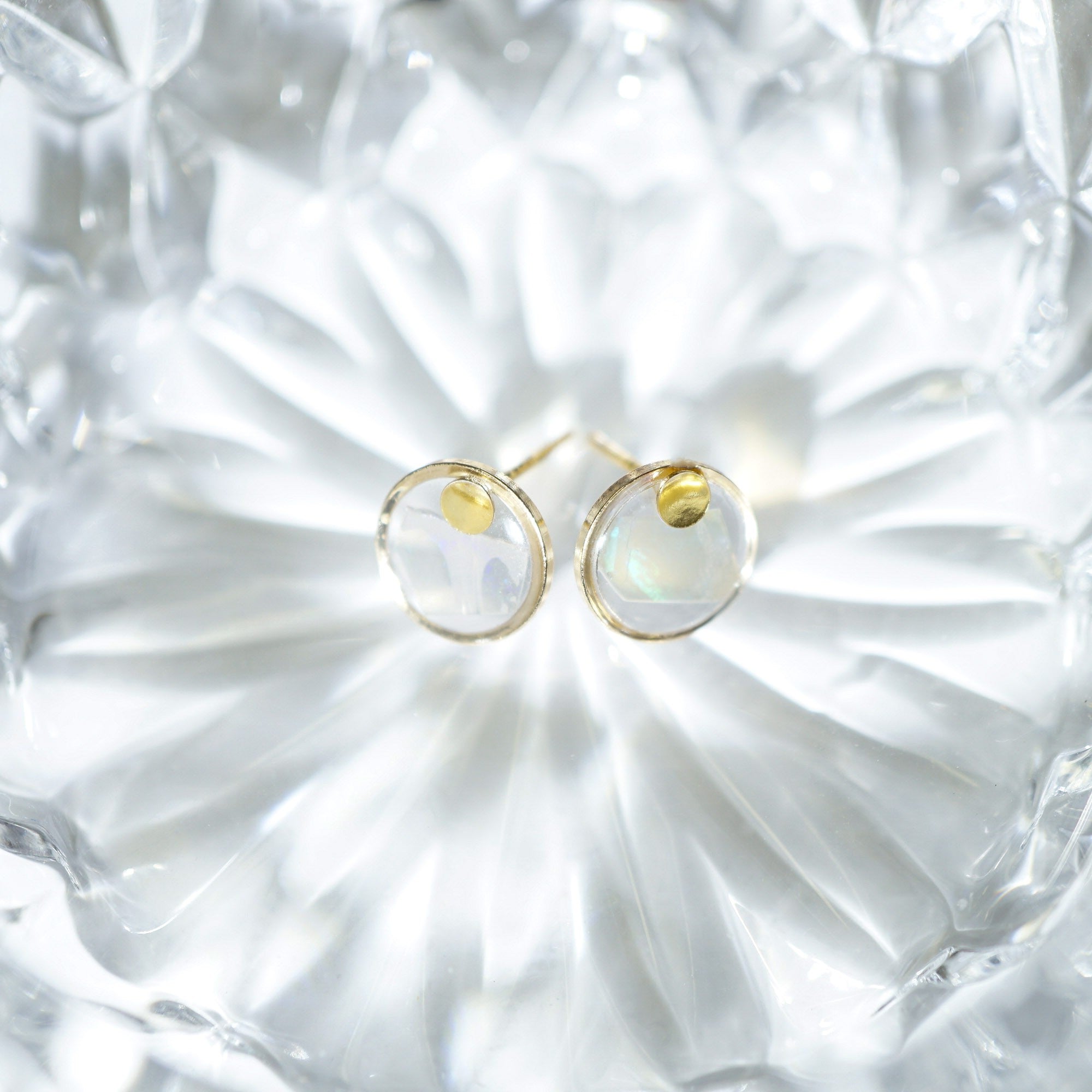 A pair of gold stud earrings with oval milky white iridescent cabochon gemstones, displayed on a textured clear glass dish.