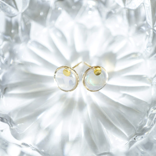 A pair of gold stud earrings with oval milky white iridescent cabochon gemstones, displayed on a textured clear glass dish.