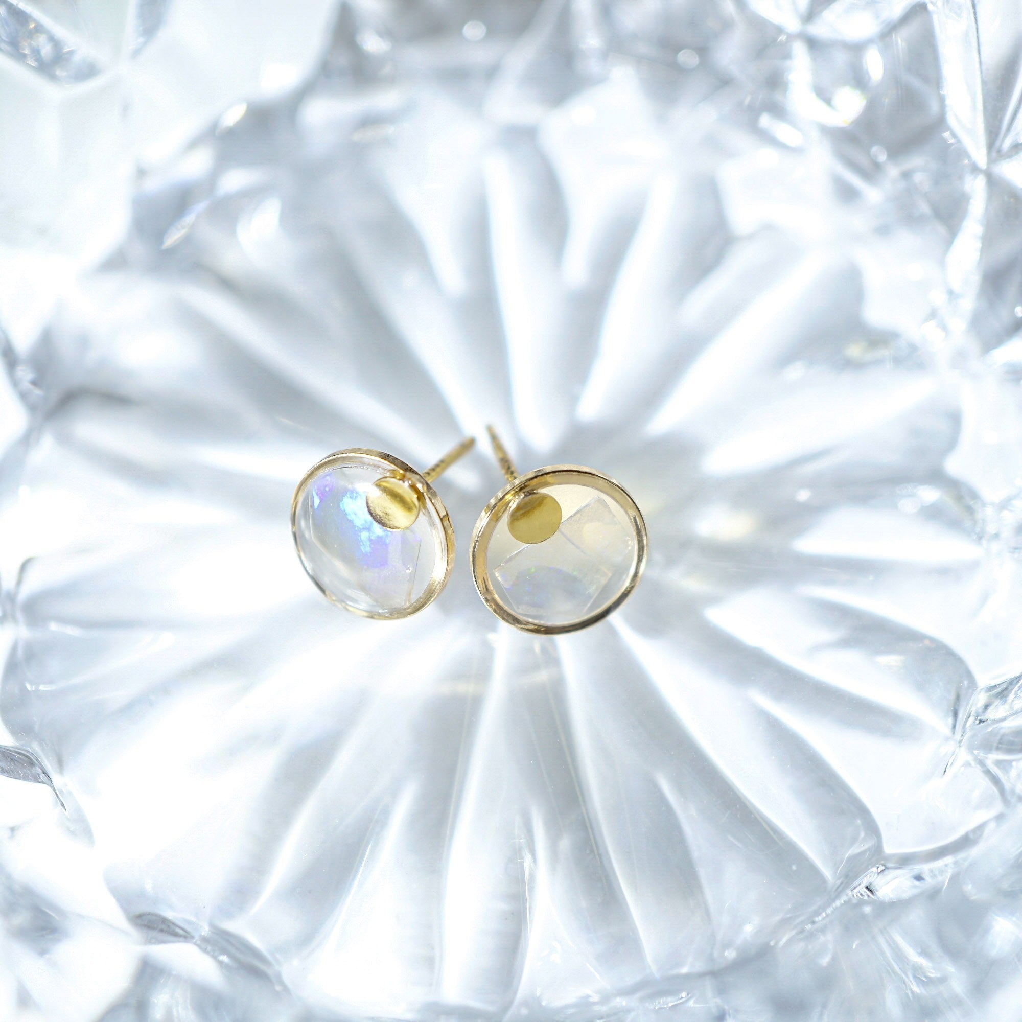 A pair of gold stud earrings with oval iridescent cabochon gemstones, displayed on a textured, translucent white glass dish.