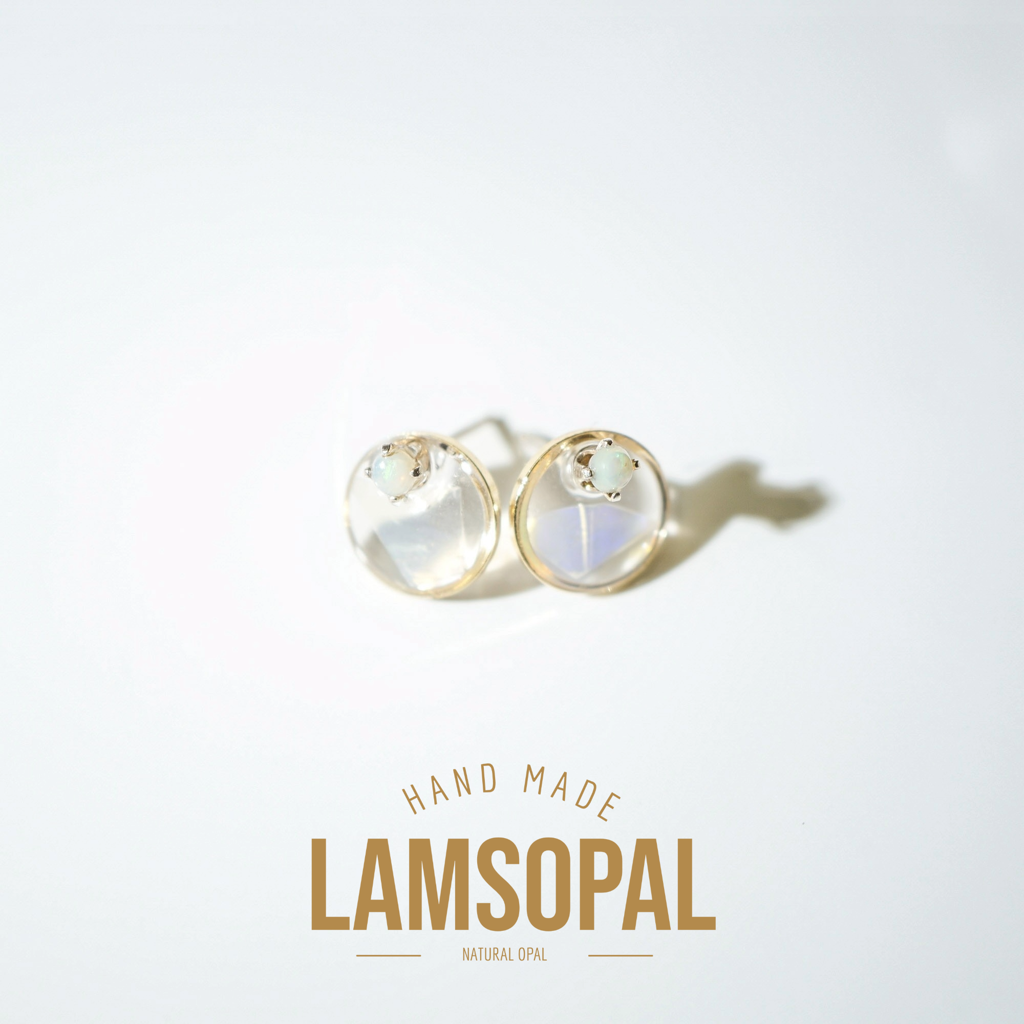 A pair of gold stud earrings with round iridescent opal cabochons, displayed on a clean white background with a soft shadow. Below the earrings, the text 'HAND MADE LAMSOPAL NATURAL OPAL' is printed in gold.