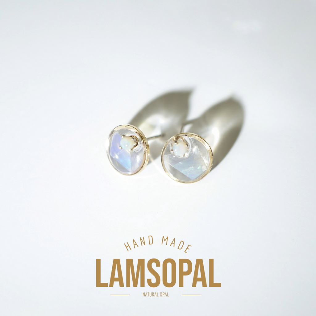 A pair of gold stud earrings with round, milky white cabochon gemstones, displayed on a clean white background with a soft shadow. Below the earrings, the text 'HAND MADE LAMSOPAL NATURAL OPAL' is printed in gold.