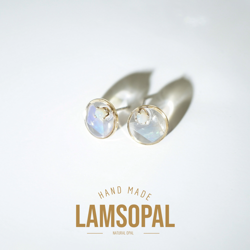 A pair of gold stud earrings with round, milky white cabochon gemstones, displayed on a clean white background with a soft shadow. Below the earrings, the text 'HAND MADE LAMSOPAL NATURAL OPAL' is printed in gold.