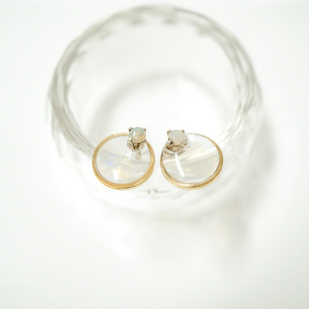 A pair of gold hoop earrings with oval-cut milky white gemstones, displayed on a clear glass dish against a bright white background.