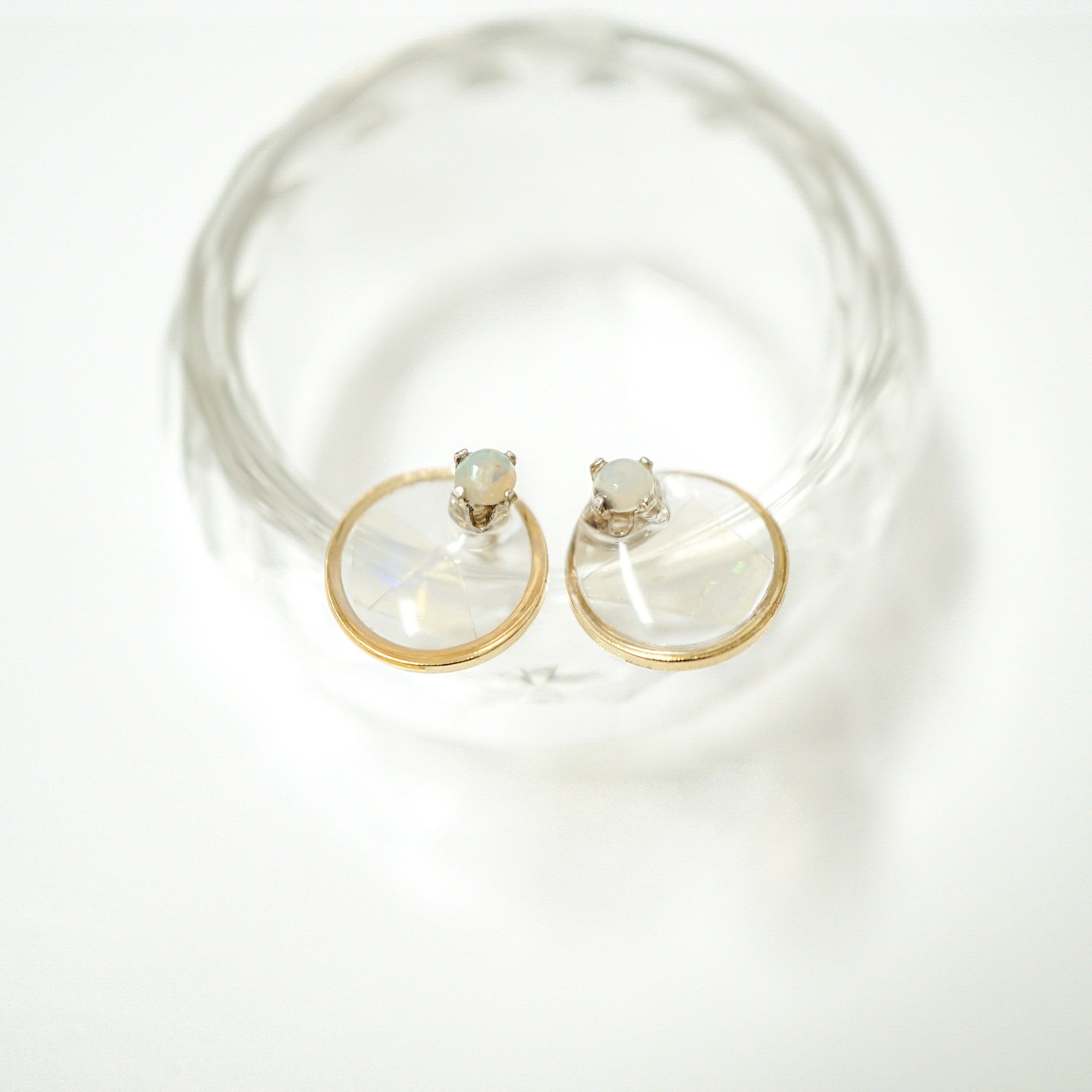 A pair of gold hoop earrings with oval-cut milky white gemstones, displayed on a clear glass dish against a bright white background.