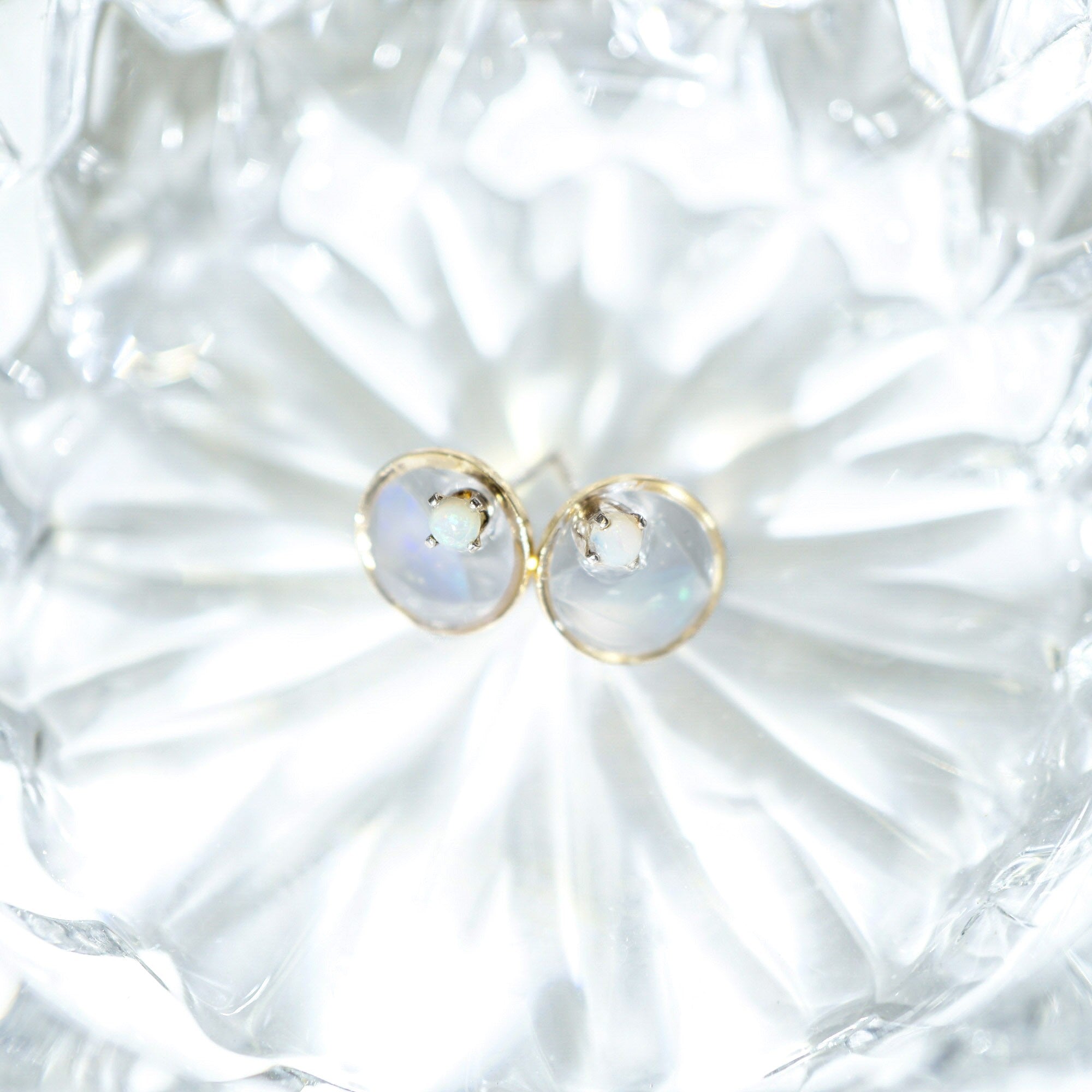 A pair of gold stud earrings with oval milky white cabochon gemstones, displayed on a textured clear glass dish.