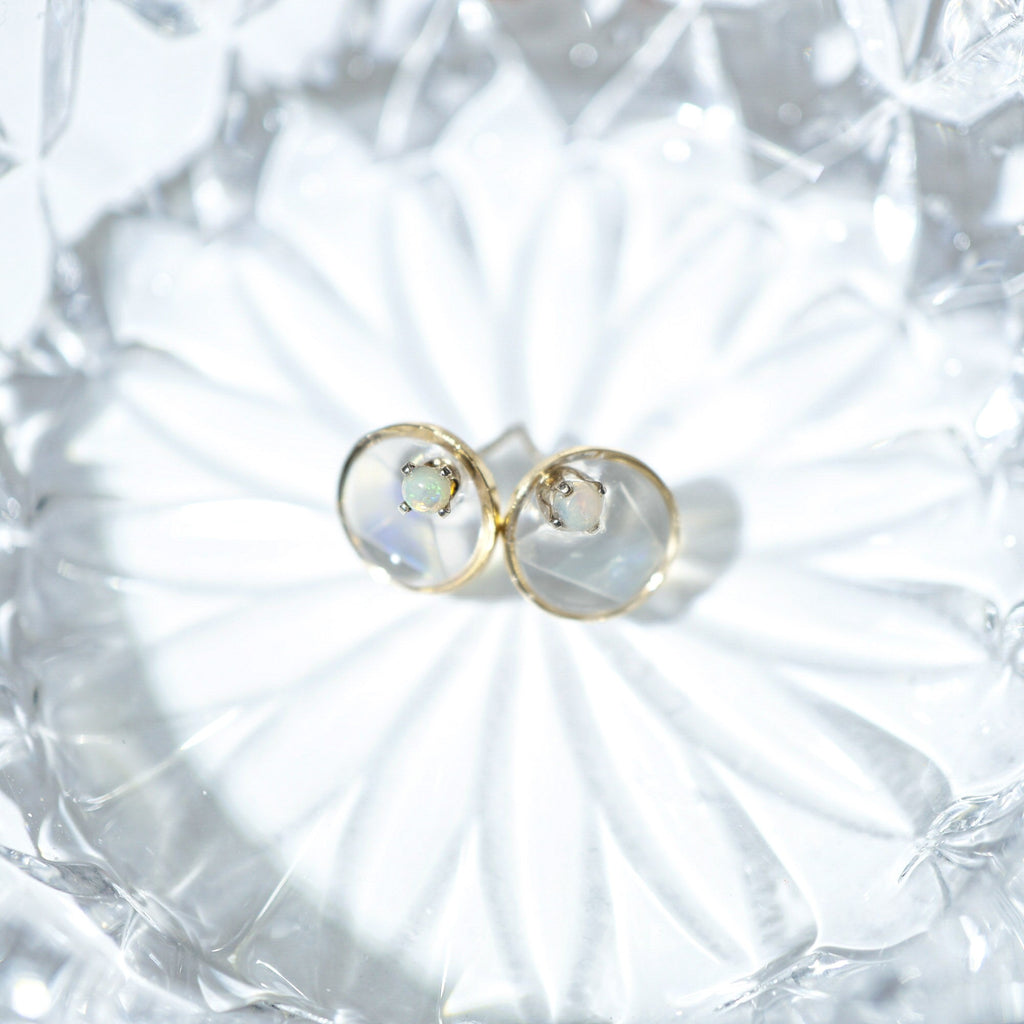 A pair of gold hoop stud earrings with small iridescent cabochon gemstones, displayed on a textured clear glass dish.