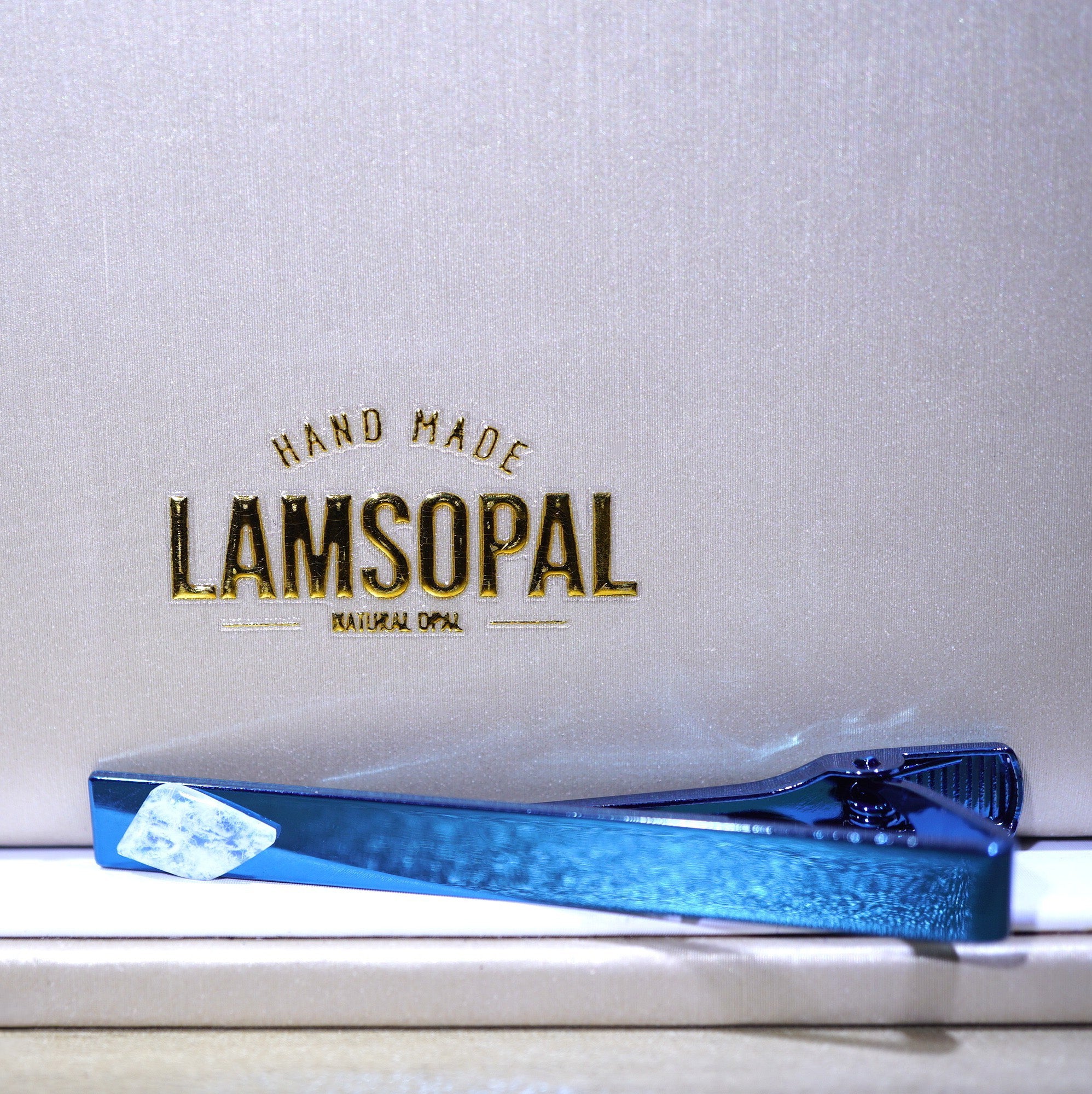 A blue glitter tie clip resting on a white surface in front of a textured light gray background with gold embossed text reading 'HAND MADE LAMSOPAL NATURAL OPAL'.