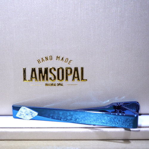 A blue glitter tie clip resting on a white surface in front of a textured light gray background with gold embossed text reading 'HAND MADE LAMSOPAL NATURAL OPAL'.