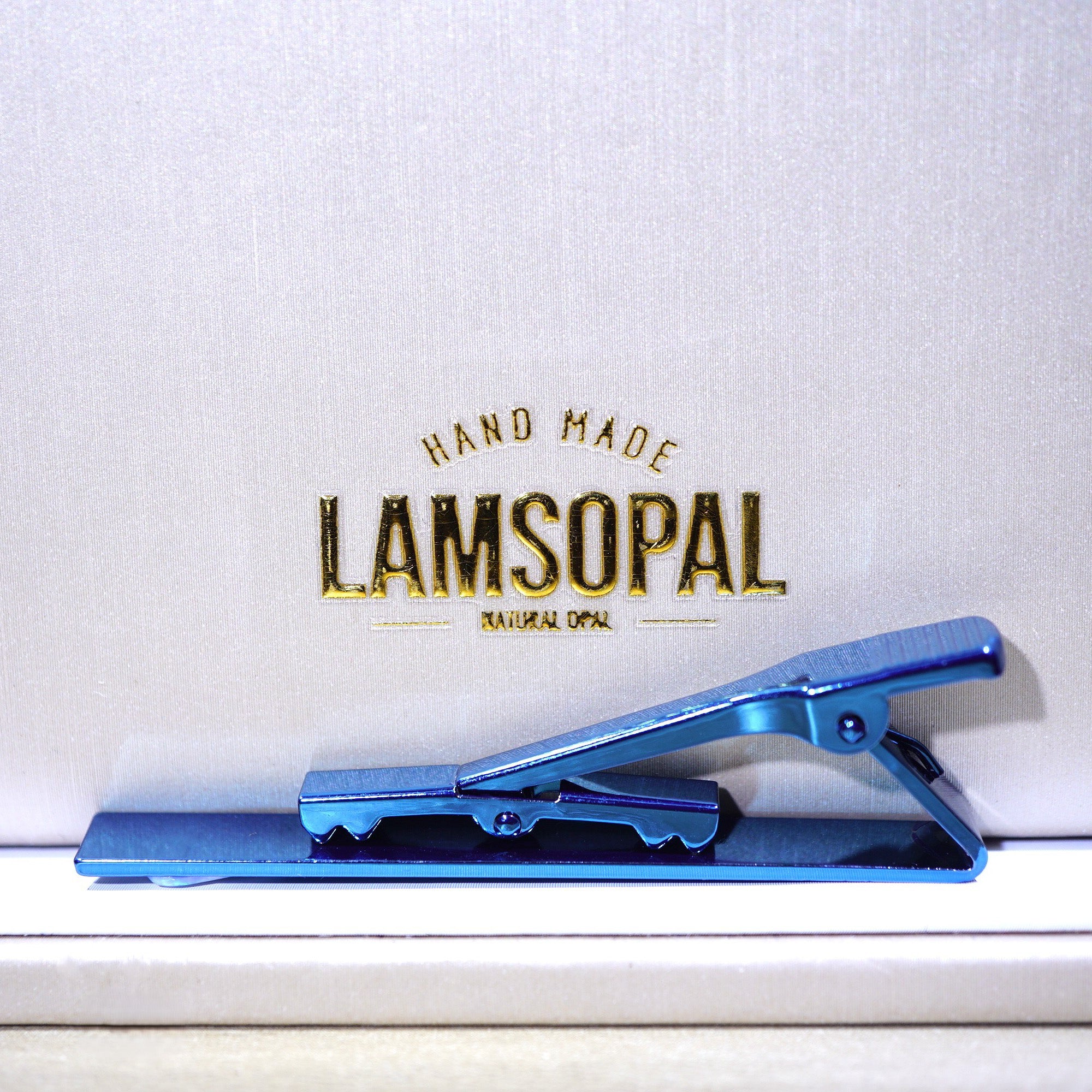 A metallic blue clip-style accessory resting on a white surface in front of a white textured background with gold text reading 'HAND MADE LAMSOPAL NATURAL OPAL'.
