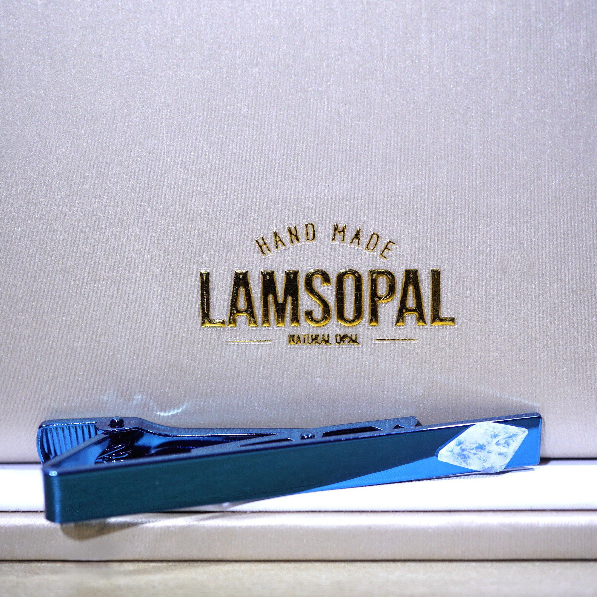 A blue Lamsopal tie clip placed on a white surface in front of a textured white background with gold 'LAMSOPAL' branding.