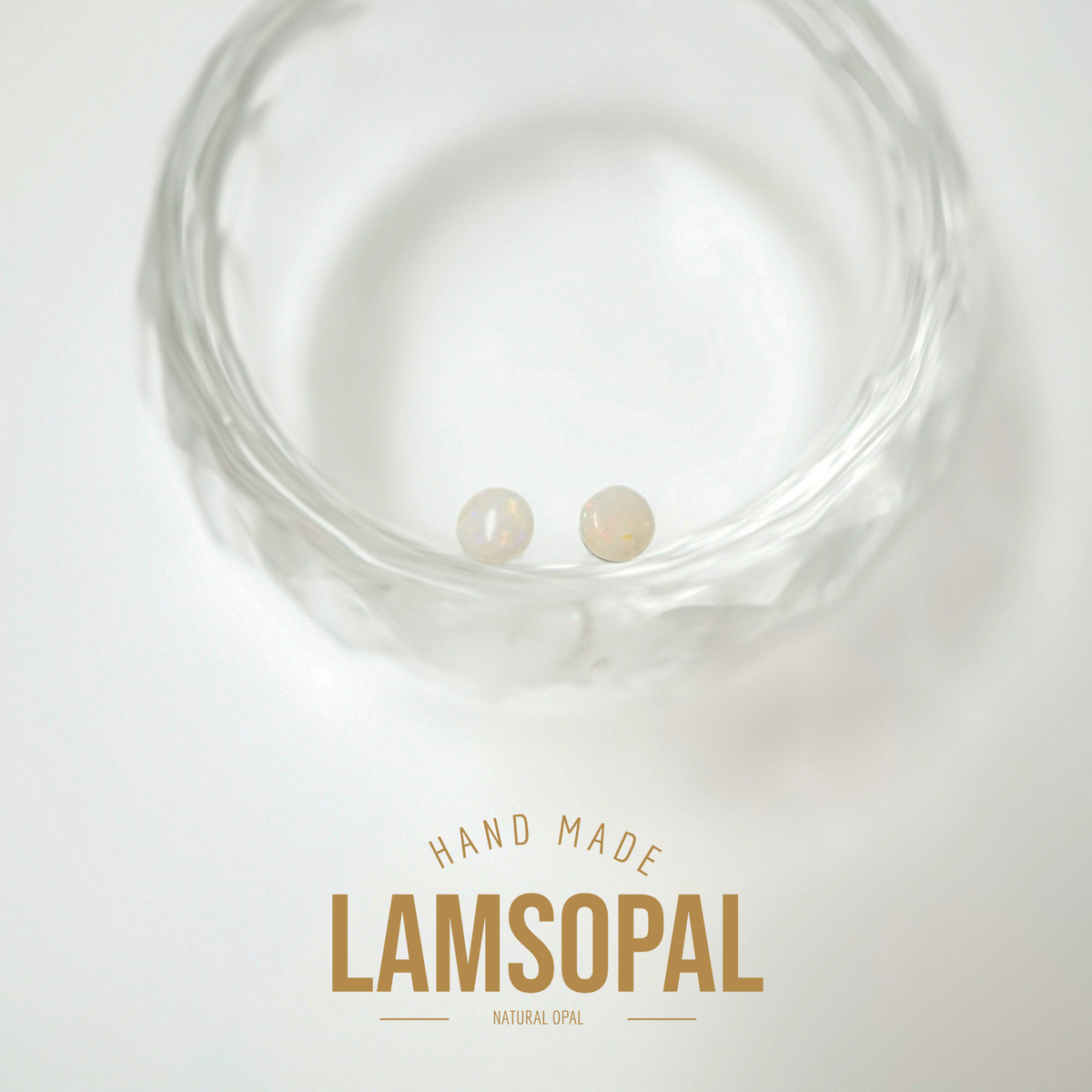 Two small natural opal gemstones displayed inside a clear textured glass bowl, with a text overlay at the bottom reading 'HAND MADE LAMSOPAL NATURAL OPAL' on a white background.