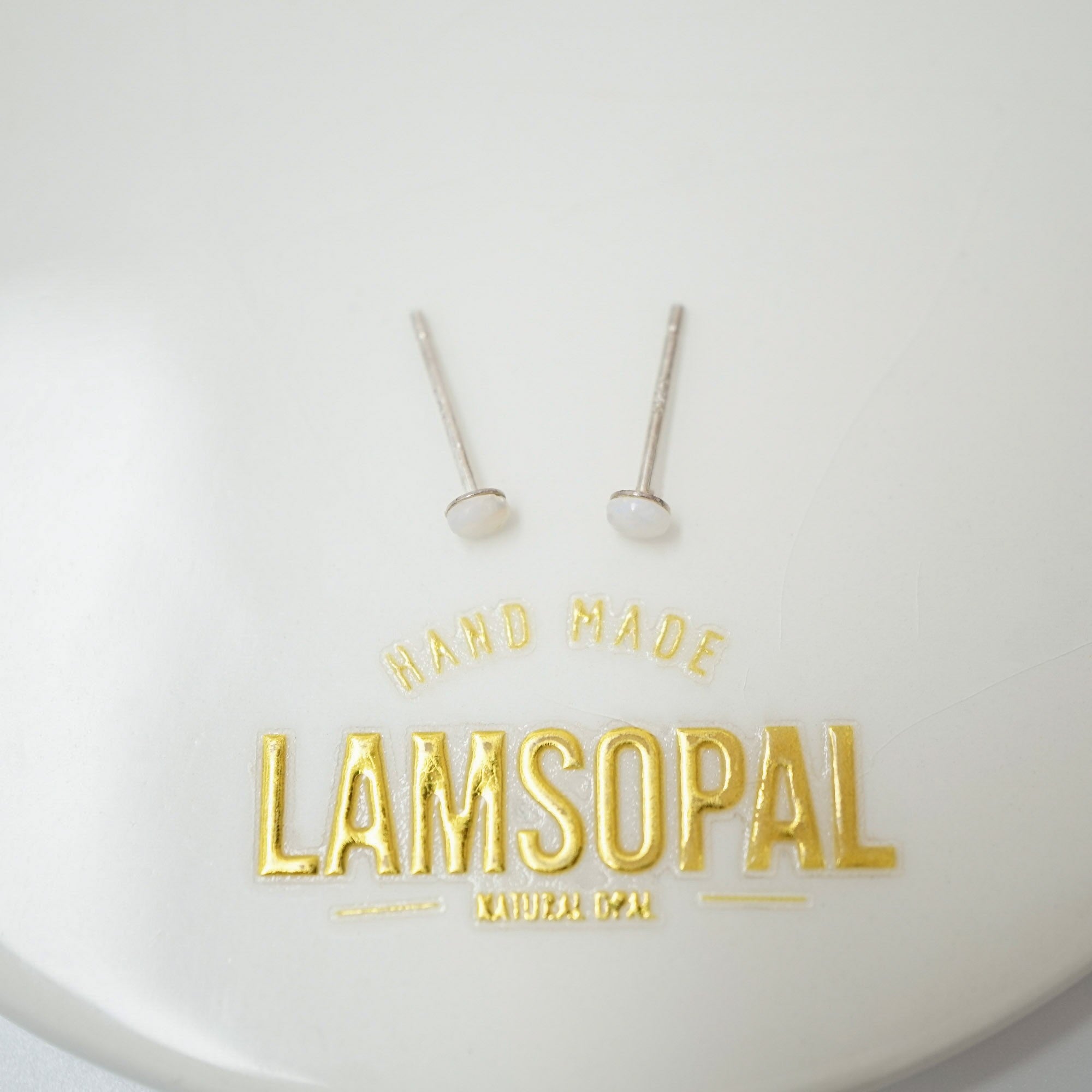 A pair of small silver-toned stud earrings with white oval inlays, displayed on a white circular surface with gold foil branding that reads 'HAND MADE LAMSOPAL NATURAL ICP'.