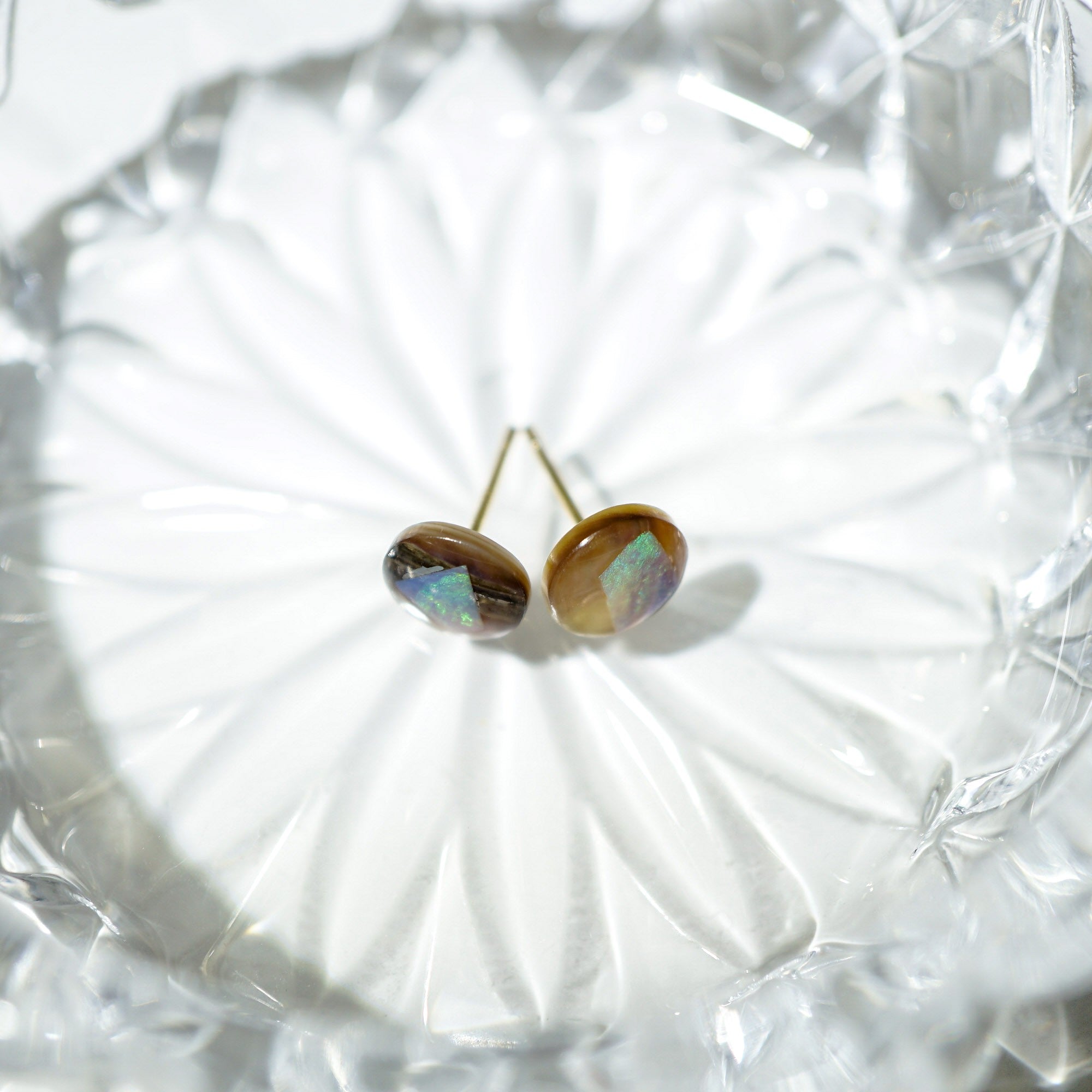 A pair of stud opal earrings with iridescent brown and blue-green stones on gold posts, displayed on a clear faceted glass dish.