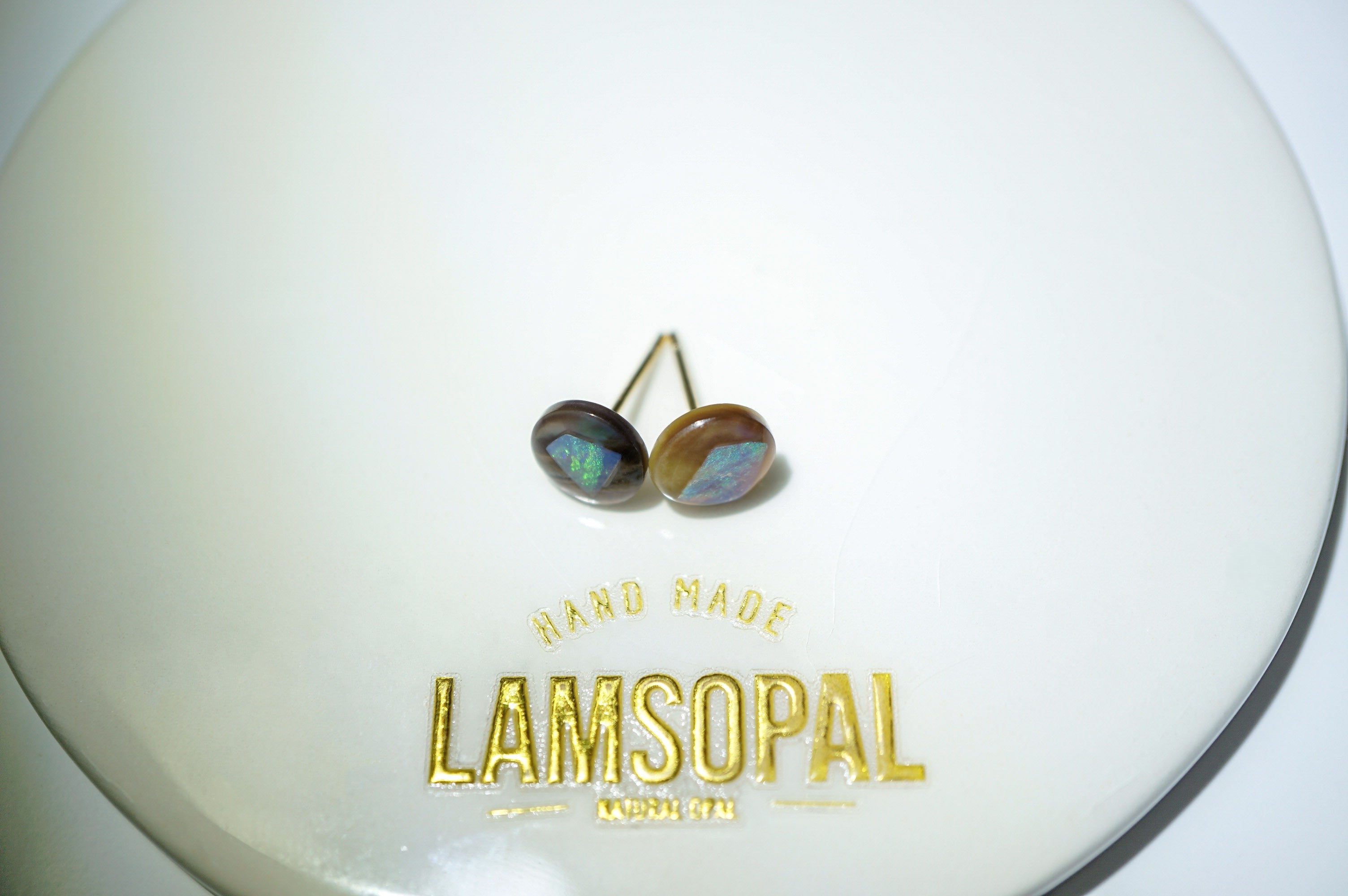 A pair of handmade Lamsopal stud earrings with iridescent blue-green and brown cabochon opal earrings set in gold, displayed on a white circular backing card with gold foil branding.