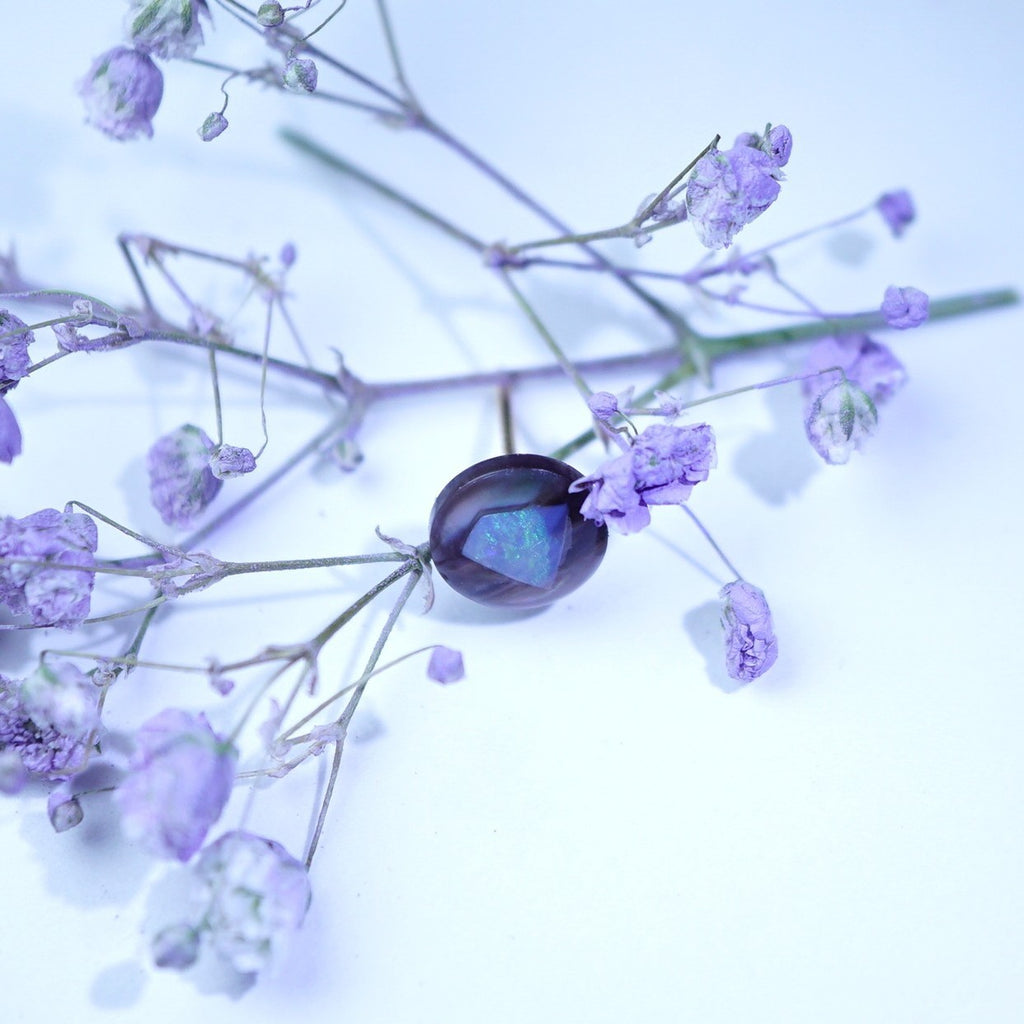 A polished oval gemstone with a deep purple body and a blue iridescent sheen, resting on delicate light purple dried flowers against a pale blue-white background.