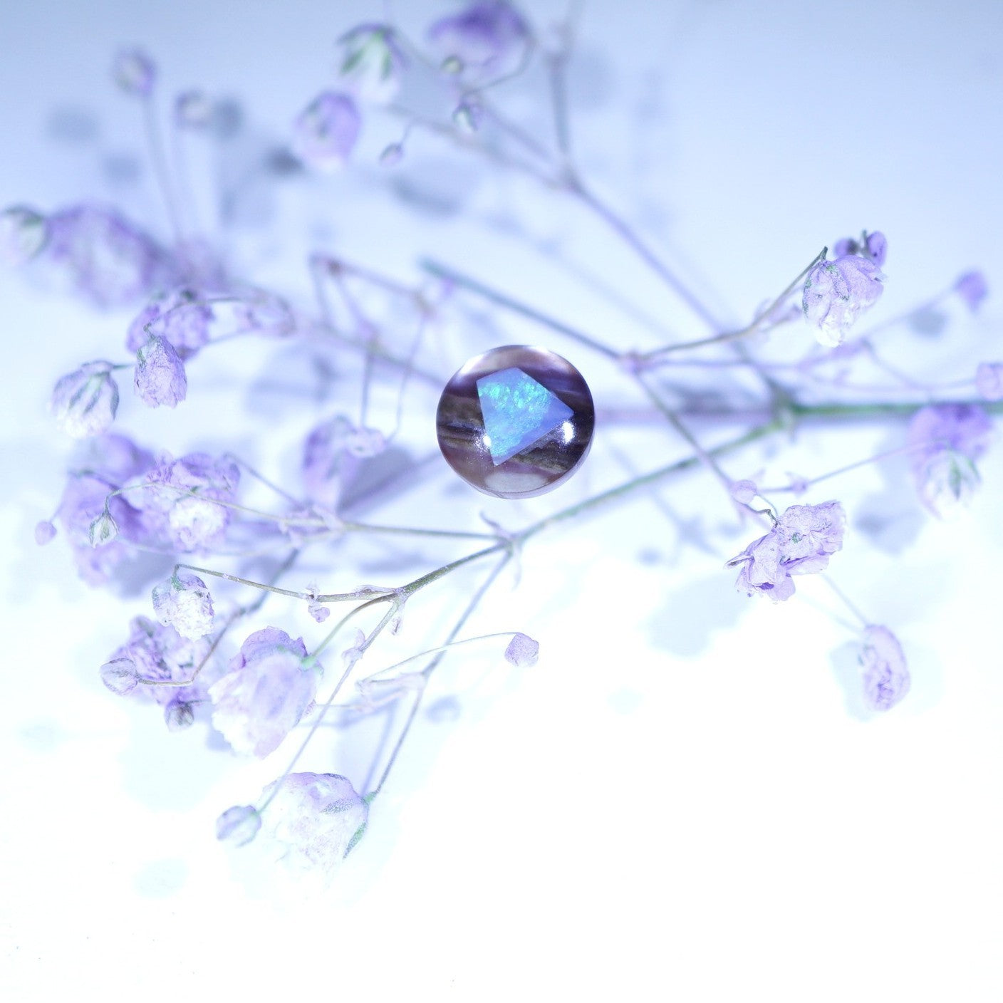 A faceted iridescent gemstone with a blue and purple rainbow sheen, resting on delicate light purple baby's breath flowers against a bright white and pale blue background.