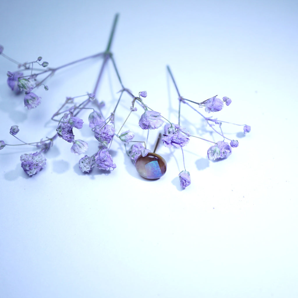 A single round cabochon gemstone with a brownish body and iridescent blue-purple flashes, set in a thin metal prong setting and surrounded by dried lavender baby's breath flowers on a light blue-white background.