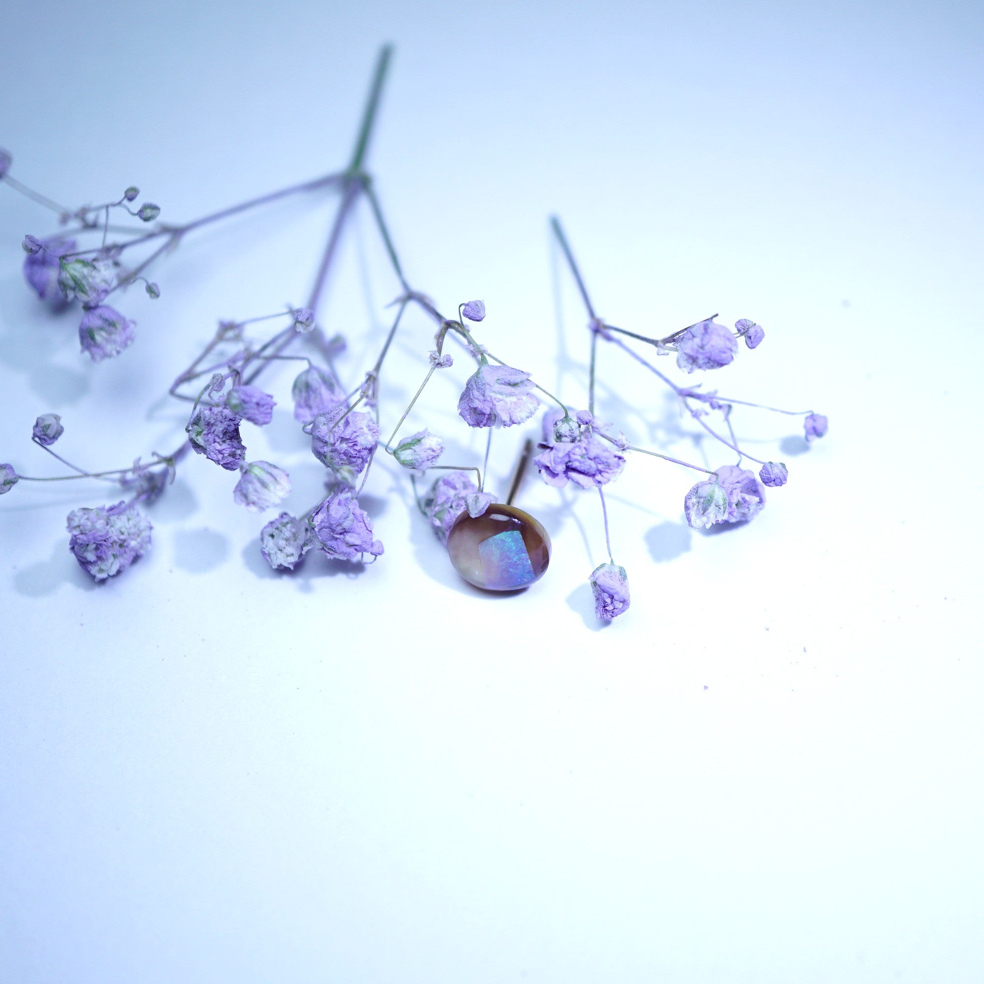A single round cabochon gemstone with a brownish body and iridescent blue-purple flashes, set in a thin metal prong setting and surrounded by dried lavender baby's breath flowers on a light blue-white background.