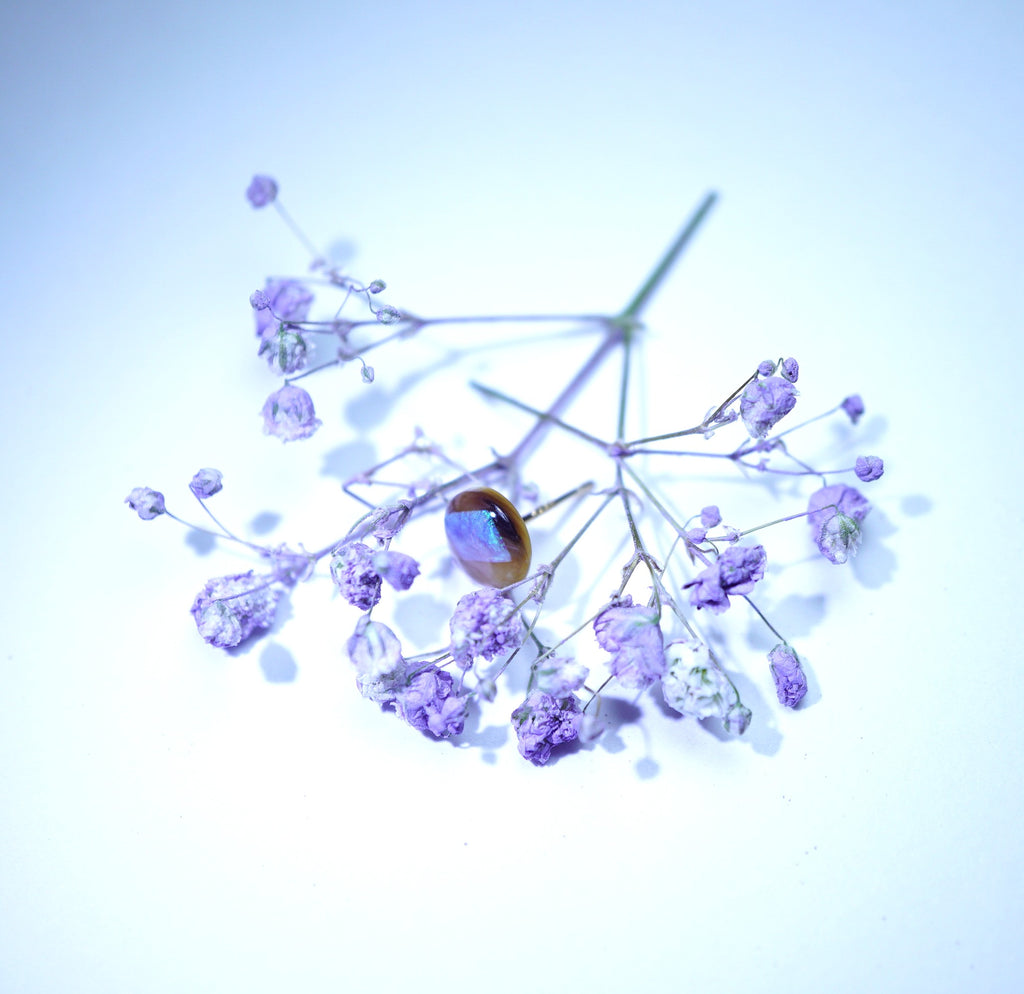 A dried floral arrangement with small lavender-purple flowers on thin stems, featuring a prominent oval iridescent brown jewel, photographed against a pale blue-white background.
