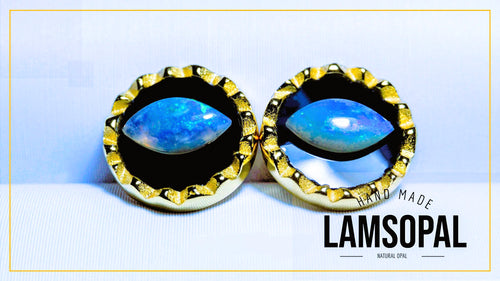 A pair of handmade Lamsopal earrings featuring natural blue opal stones in circular settings with textured gold borders, displayed on a light background with a gold border and brand text overlay.