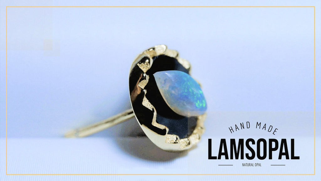 A gold-toned ring with a large, irregularly shaped natural blue opal gemstone, shown on a light blue background with a gold border. The text 'HAND MADE LAMSOPAL' is visible in the bottom right corner.