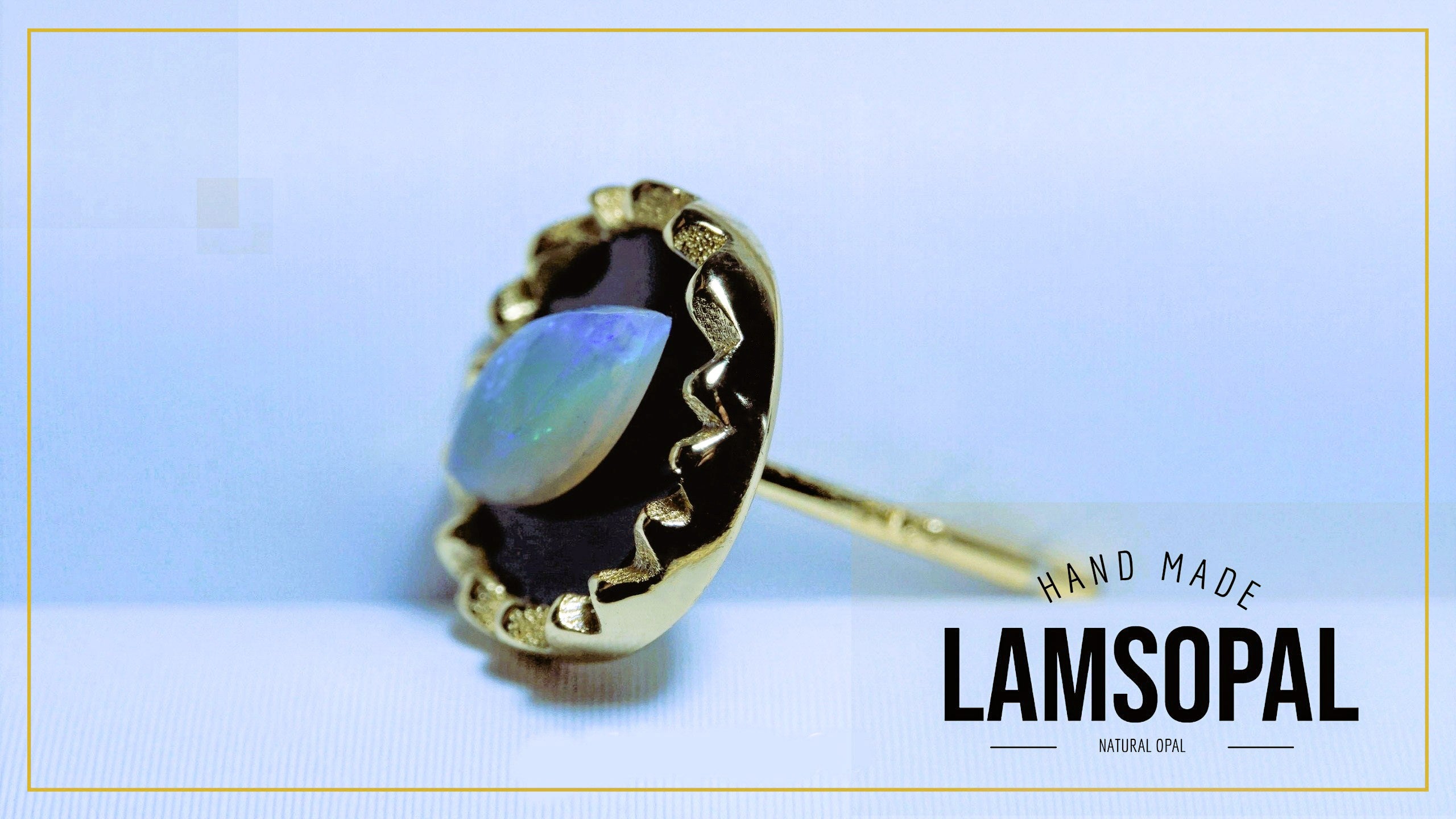 A gold ring with a natural opal gemstone, featuring a wavy bezel setting and a post, shown against a light blue background with a gold border. The text 'HAND MADE LAMSOPAL' is visible in the bottom right corner.
