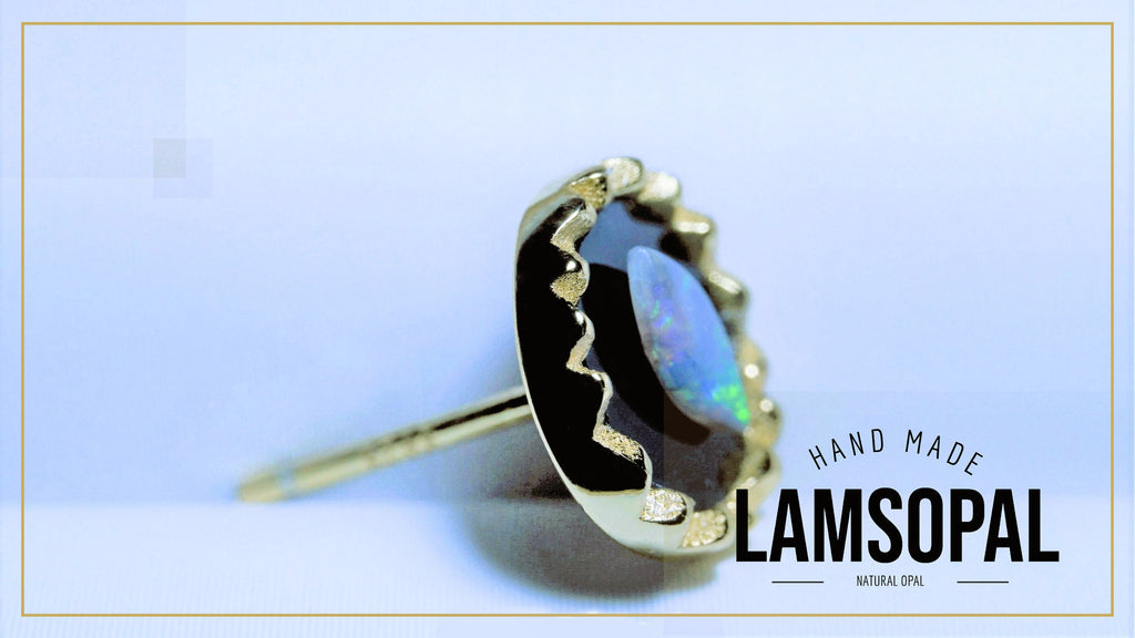 A gold ring featuring a large, irregularly shaped black opal with vibrant blue and green iridescent flashes, set in a gold bezel with a wavy gold accent line. The ring is displayed on a light blue background with a gold border, and a text overlay on the bottom right reads 'HAND MADE LAMSOPAL NATURAL OPAL'.
