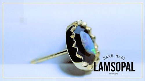 A gold ring featuring a large, irregularly shaped black opal with vibrant blue and green iridescent flashes, set in a gold bezel with a wavy gold accent line. The ring is displayed on a light blue background with a gold border, and a text overlay on the bottom right reads 'HAND MADE LAMSOPAL NATURAL OPAL'.