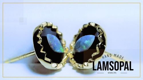 A pair of gold-toned lapel pins with oval black opal stones, featuring a wavy gold bezel setting and a post with a butterfly back. The image has a light blue background with a gold border, and a text overlay on the bottom right reads 'HAND MADE LAMSOPAL NATURAL OPAL'.