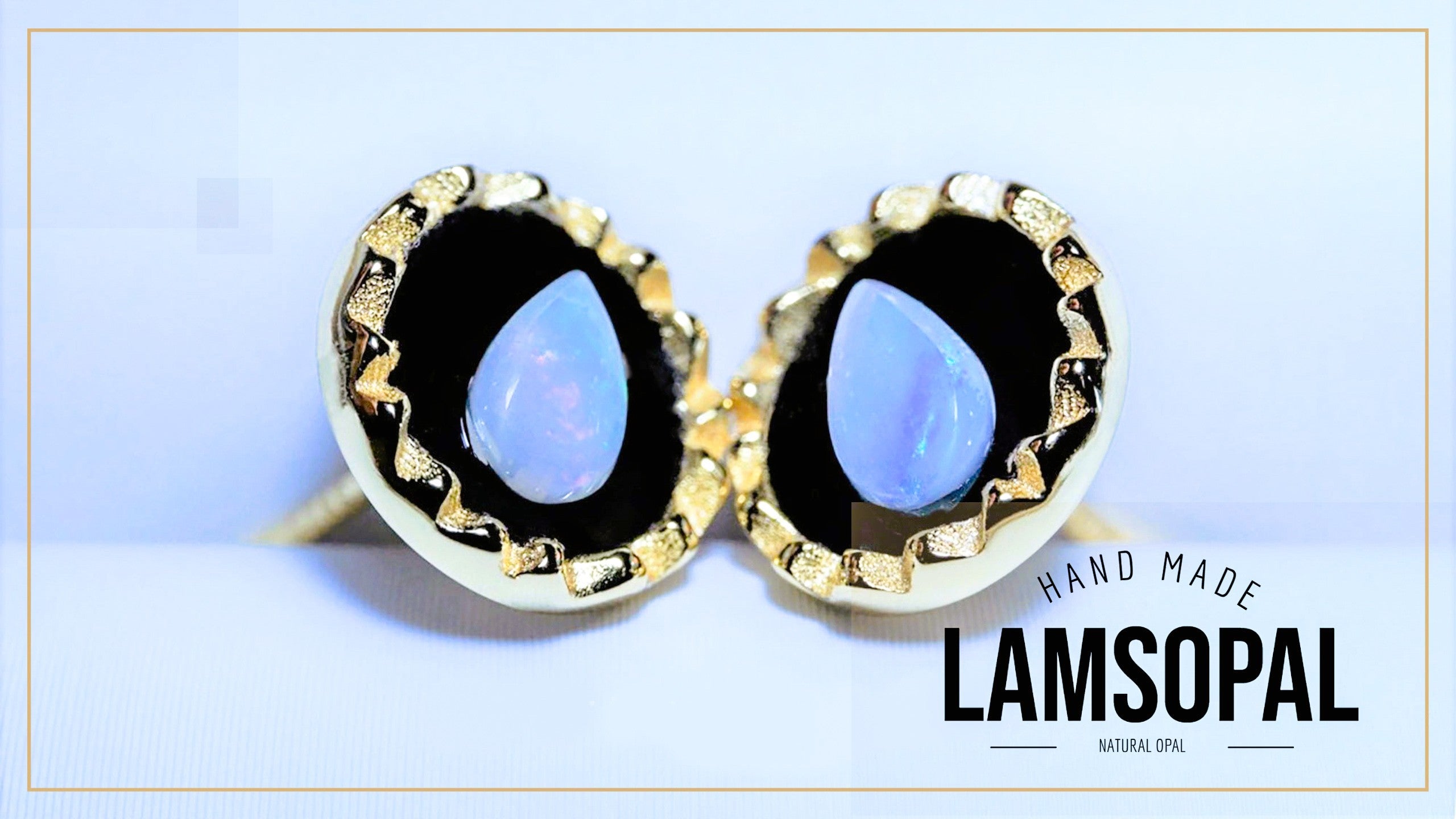 A pair of gold-toned stud earrings with teardrop-shaped light blue opal centers, set in black bezel settings with a textured gold border. The image features a 'HAND MADE LAMSOPAL NATURAL OPAL' text overlay in the bottom right corner.