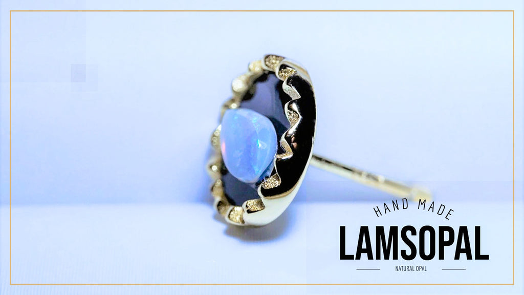 A handmade Lamsopal ring featuring a natural opal gemstone with iridescent blue, purple, and green flashes, set in a gold-toned bezel with a black inlay. The ring is displayed on a light blue background with a gold border, and the text 'HAND MADE LAMSOPAL' is visible in the bottom right corner.
