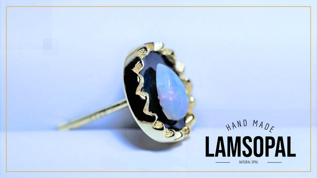 A gold-toned Lamsopal stud earring featuring a round natural opal with iridescent blue, purple, and green flashes, set in a bezel with a wavy gold border. The earring is displayed on a light blue background with a gold border, and the text 'HAND MADE LAMSOPAL' is visible in the bottom right corner.