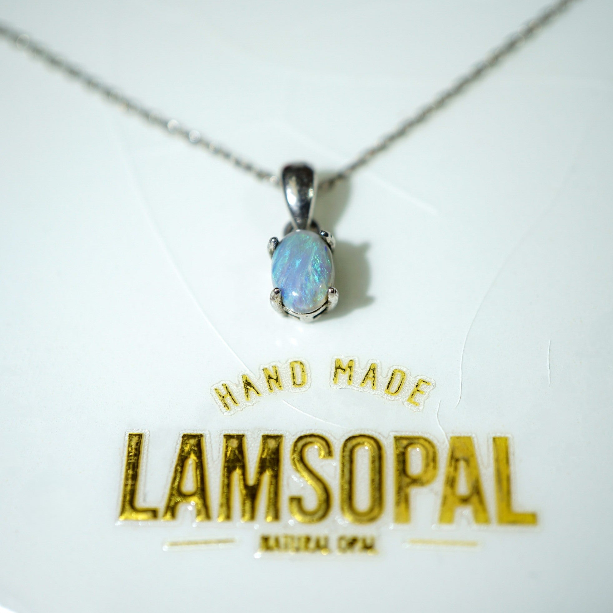 A silver pendant necklace with an oval iridescent opal stone, displayed on a white surface above a gold 'LAMSOPAL' brand logo.